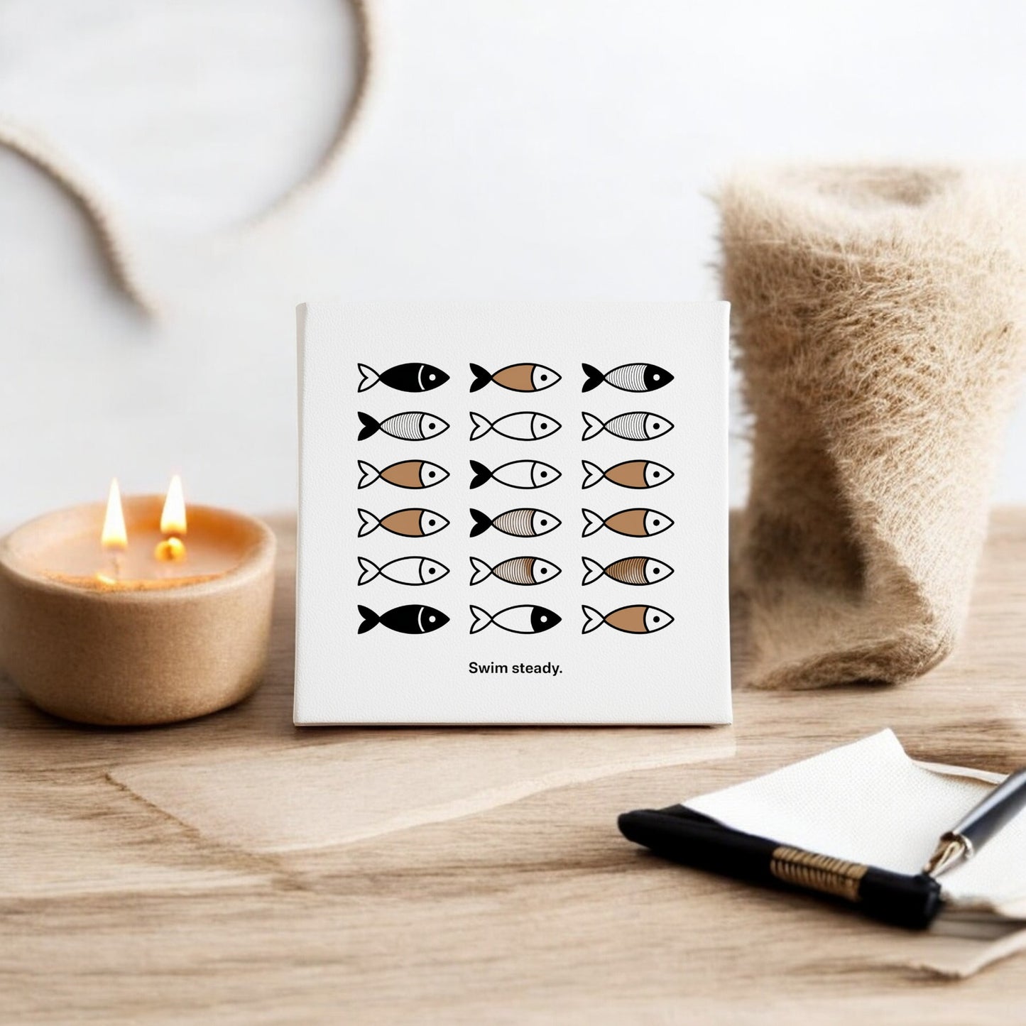 The Swim Steady Canvas by Mirabilia Boutique is an 8x8 minimalist neutral wall art, featuring rows of stylized black, white, and brown fish with the text “Swim steady.” Displayed on a wooden surface with a candle, cloth, pen, and napkin nearby.