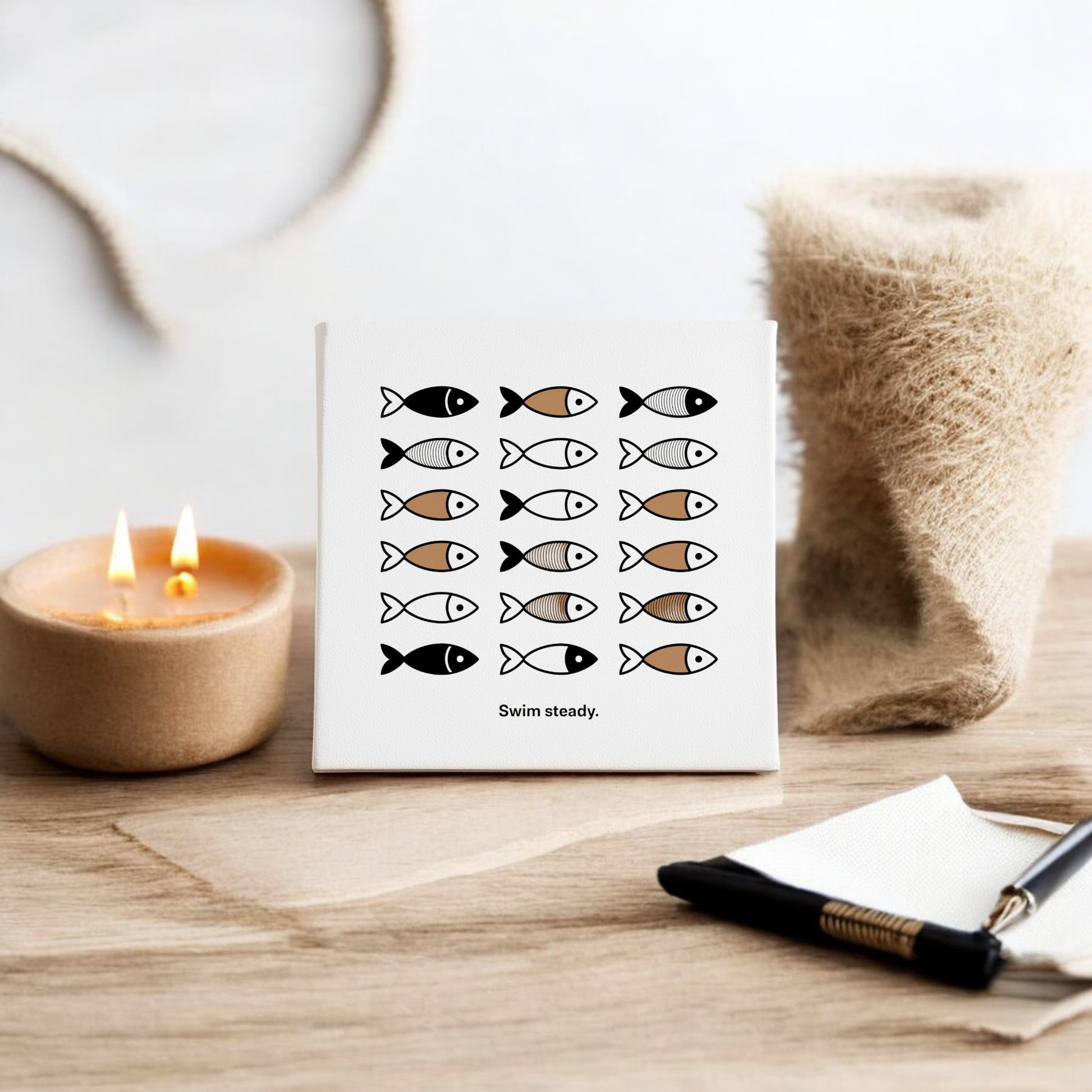 The Swim Steady Canvas by Mirabilia Boutique is an 8x8 minimalist neutral wall art, featuring rows of stylized black, white, and brown fish with the text “Swim steady.” Displayed on a wooden surface with a candle, cloth, pen, and napkin nearby.