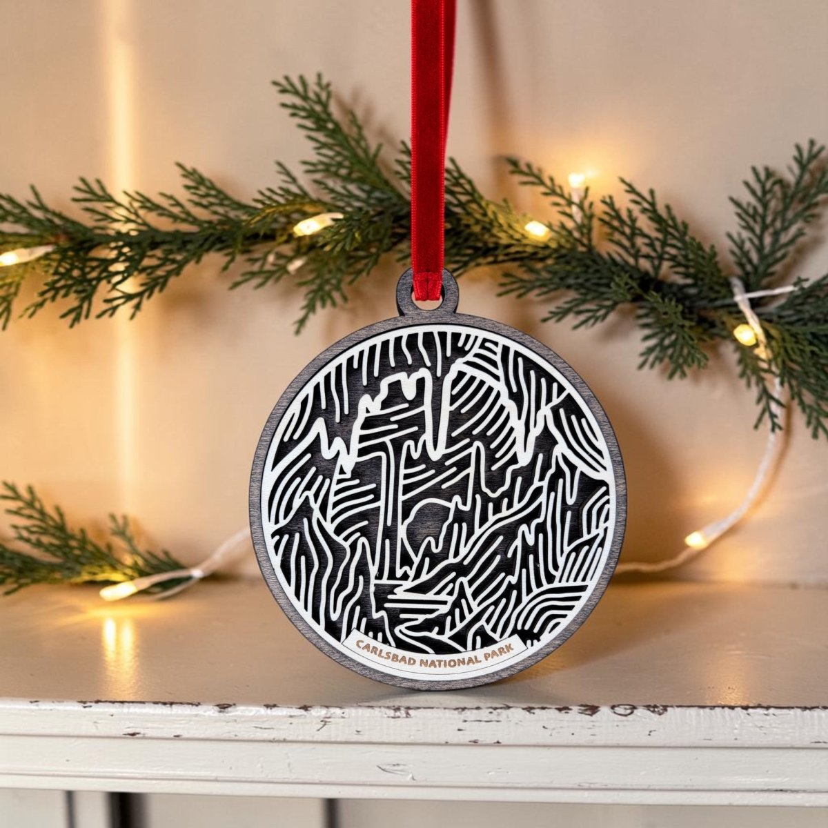 The Carlsbad Caverns National Park Christmas Ornament by Mirabilia Boutique is a handcrafted wooden ornament showcasing a bold black and white cave design and the words "Carlsbad National Park." Hanging from a red ribbon with pine branches and string lights in the background, this unique piece is the perfect way to celebrate your love for adventure.