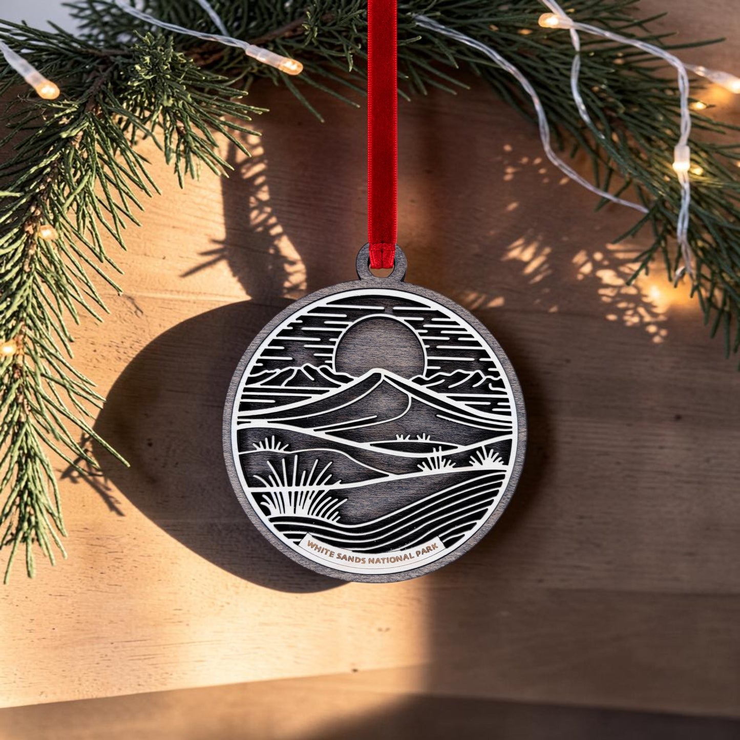 The White Sands National Park Line Art Christmas Ornament from Mirabilia Boutique features a landscape design of the shimmering gypsum dunes on an oval metal ornament, hanging from a red ribbon. "White Sands National Park" is printed on the ornament, set against a background of pine branches and glowing string lights.