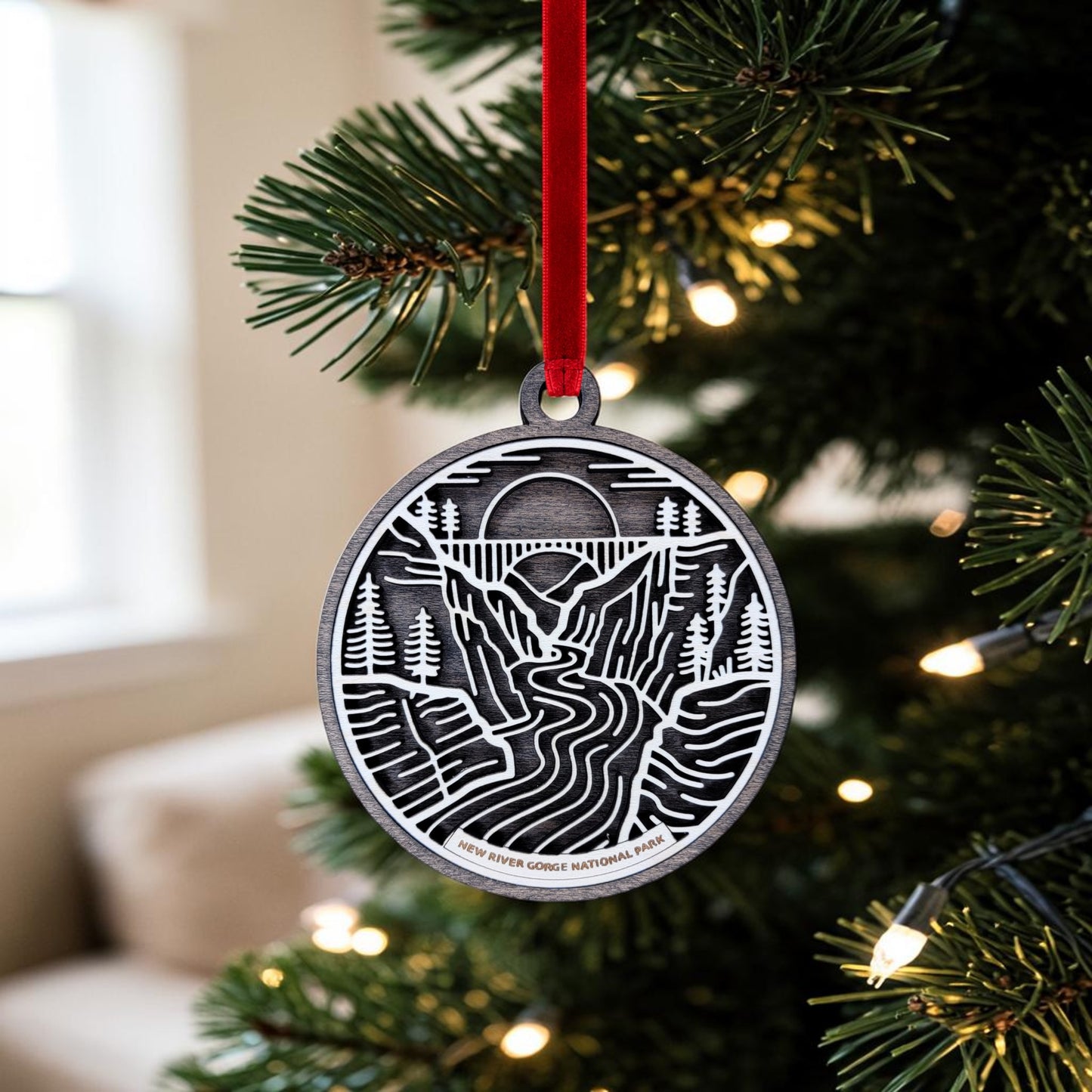 A New River Gorge National Park Line Art Christmas Ornament by Mirabilia Boutique hangs from a red ribbon on a Christmas tree with lights. This metal ornament features the New River Gorge Bridge spanning a river, framed by trees, mountains, and a sunset—capturing the beauty of New River Gorge National Park.