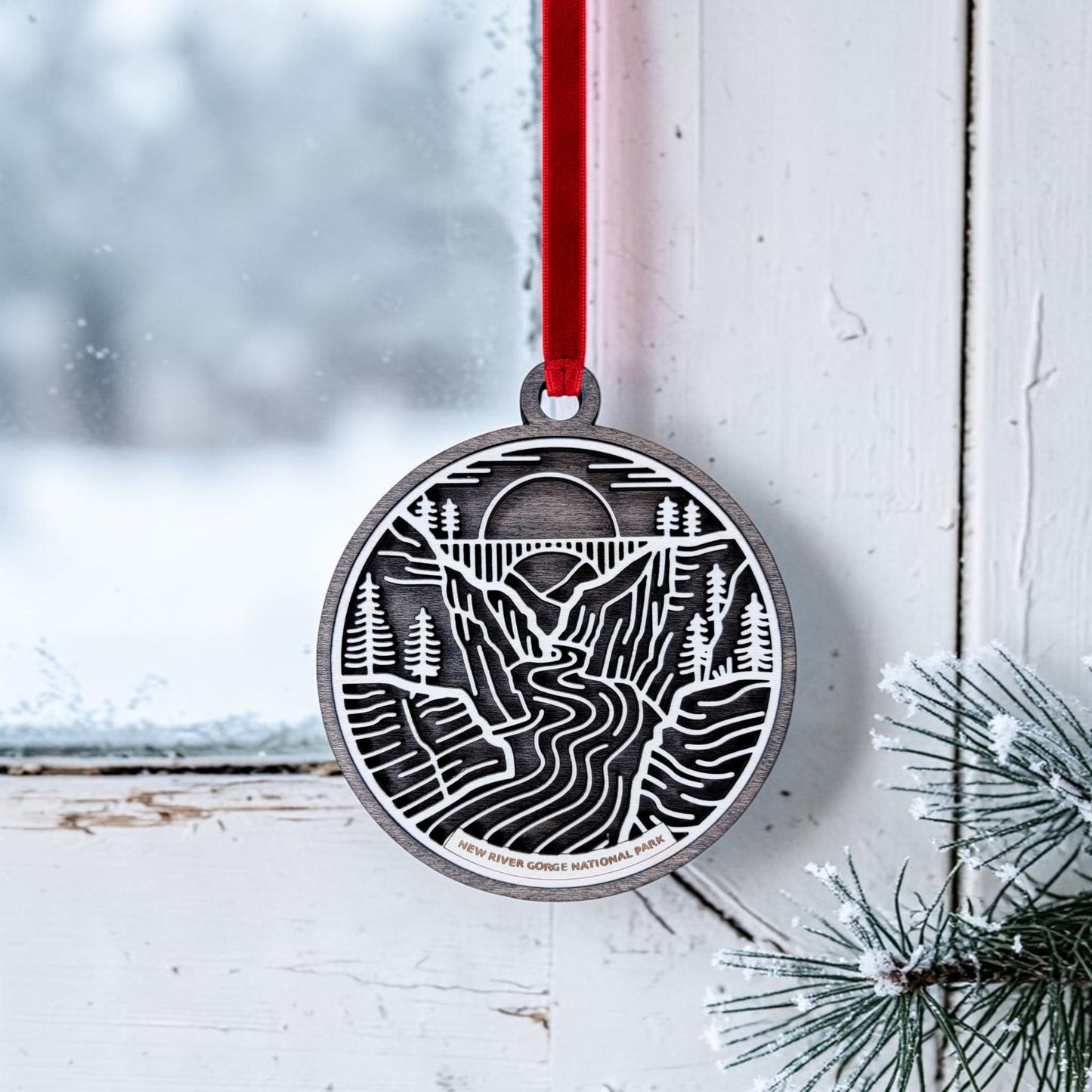 A New River Gorge National Park Line Art Christmas Ornament by Mirabilia Boutique, showcasing a river, trees, and sun design on round wood, hangs from a red ribbon in a snowy window. Pine branches dusted with snow frame the scene, capturing the tranquil beauty of this iconic park.