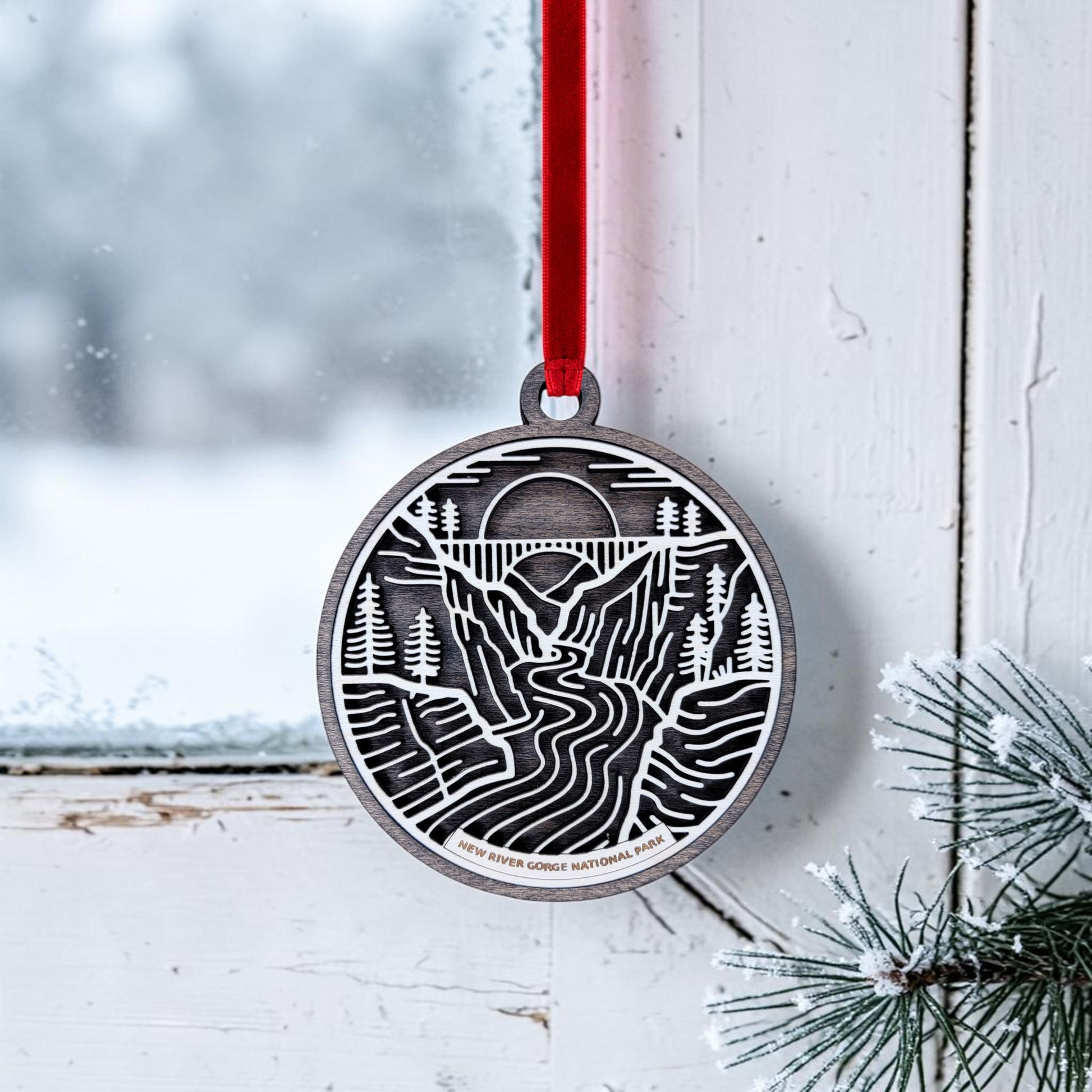 A New River Gorge National Park Line Art Christmas Ornament by Mirabilia Boutique, showcasing a river, trees, and sun design on round wood, hangs from a red ribbon in a snowy window. Pine branches dusted with snow frame the scene, capturing the tranquil beauty of this iconic park.