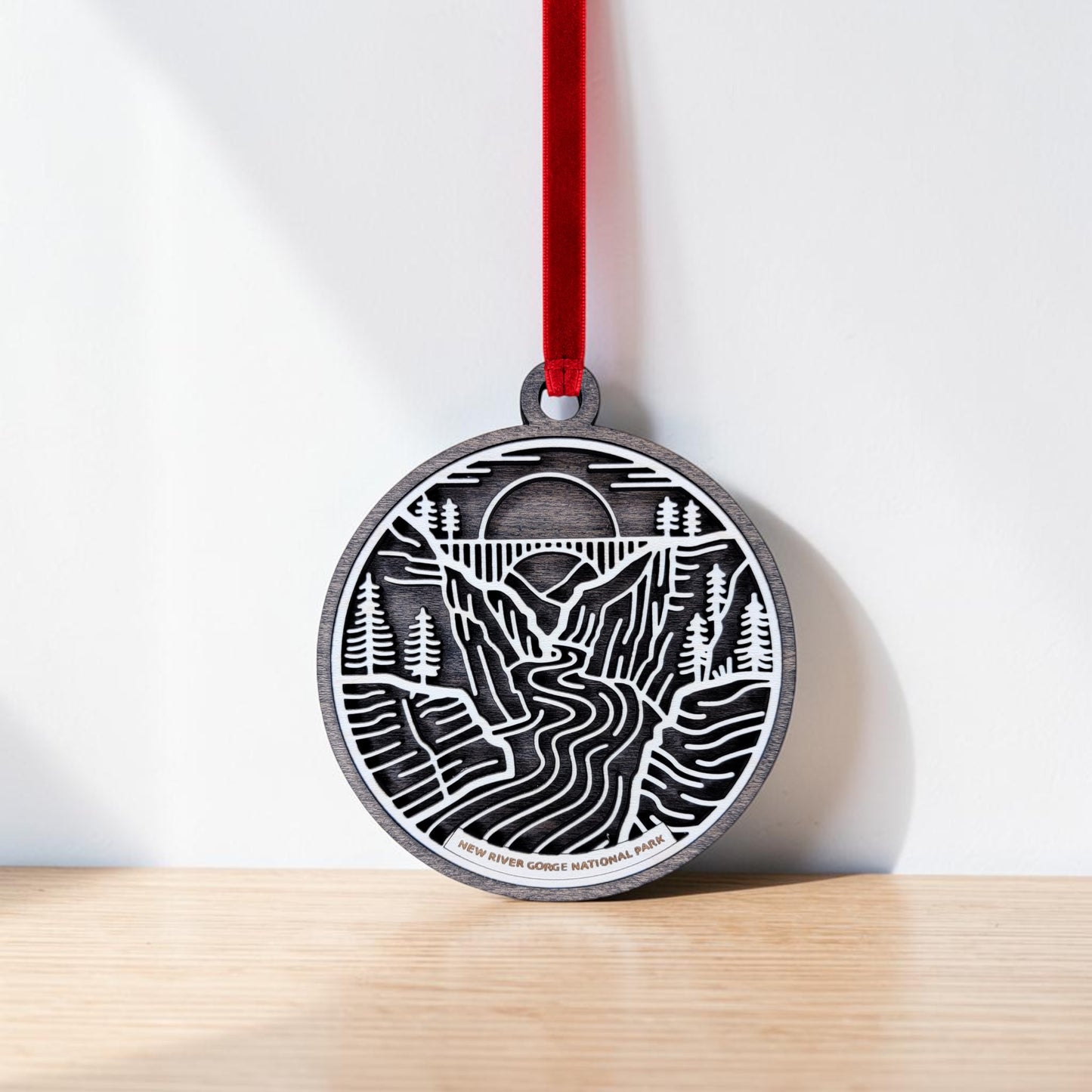 The New River Gorge National Park Line Art Christmas Ornament by Mirabilia Boutique—a round wooden piece engraved with a detailed landscape of mountains, trees, and a river—hangs from a red ribbon against a white wall and wooden surface.