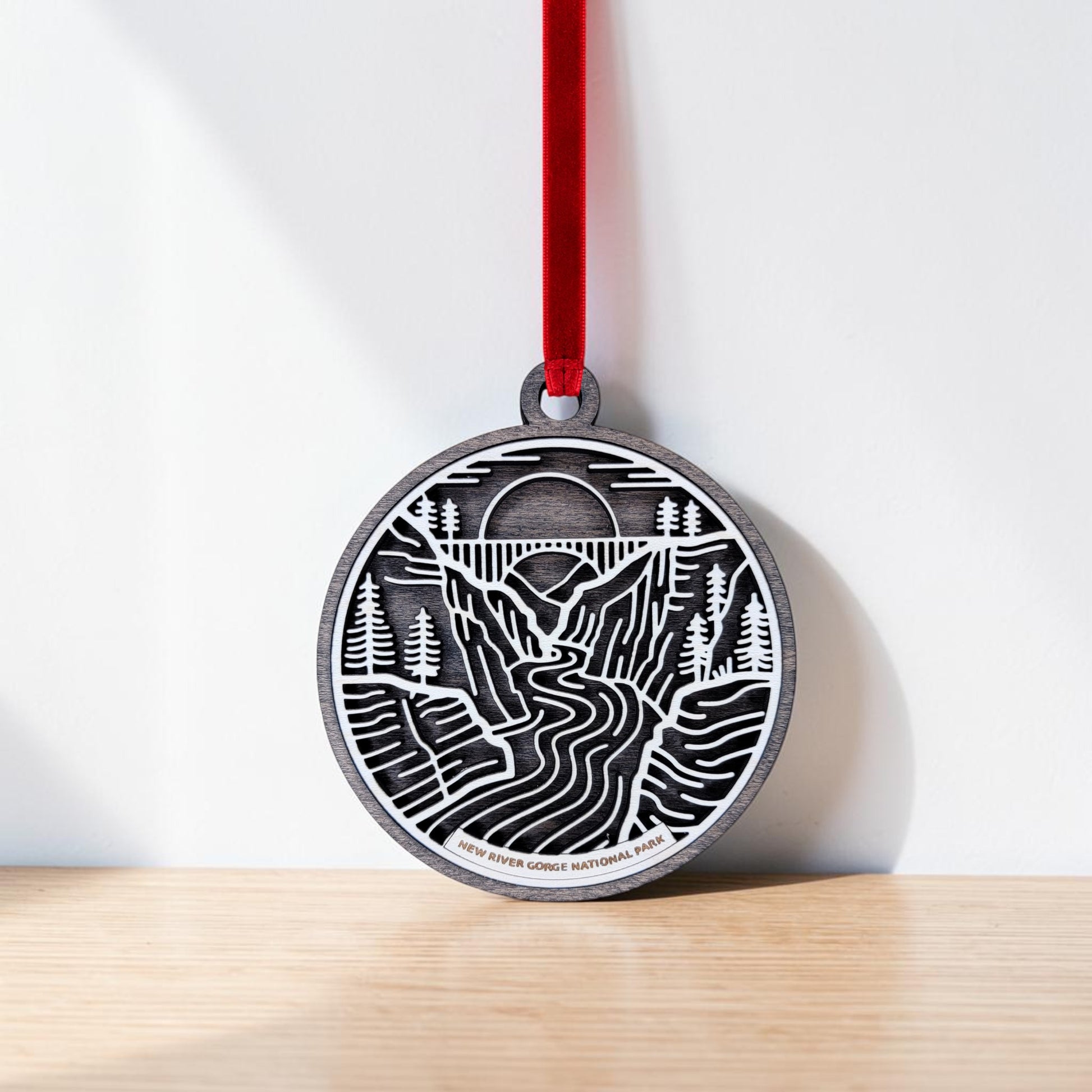 The New River Gorge National Park Line Art Christmas Ornament by Mirabilia Boutique—a round wooden piece engraved with a detailed landscape of mountains, trees, and a river—hangs from a red ribbon against a white wall and wooden surface.