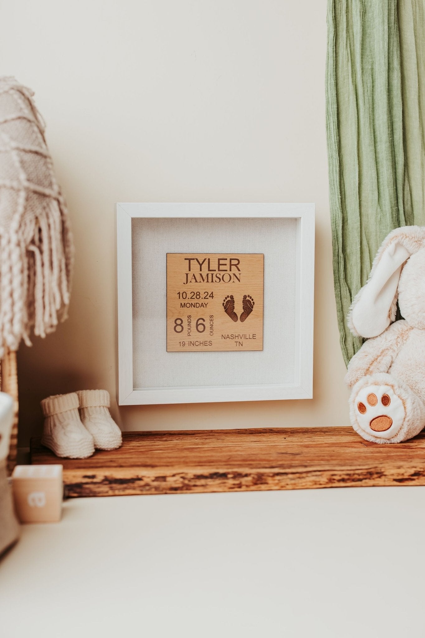 The Mirabilia Boutique Personalized Baby Birth Stats Shadowbox, a 9x9 custom newborn name sign, is displayed with small shoes, a woven blanket, green cloth, and plush bunny on a wooden shelf for a charming nursery touch.