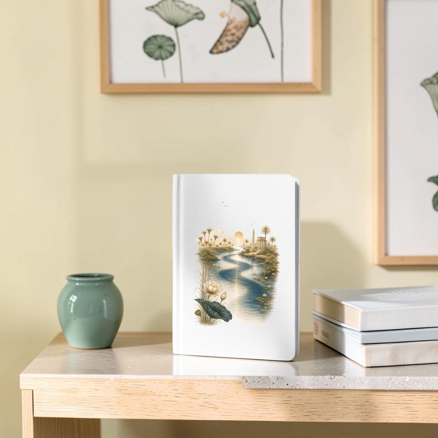 The Nile Path Journal | Ancient Egyptian Inspired Notebook by Mirabilia Boutique—a white notebook featuring a tranquil river and nature scene with delicate lotus and papyrus motifs—rests on a light wood table beside a green vase and two closed books, while framed botanical art decorates the pale yellow wall in the background.