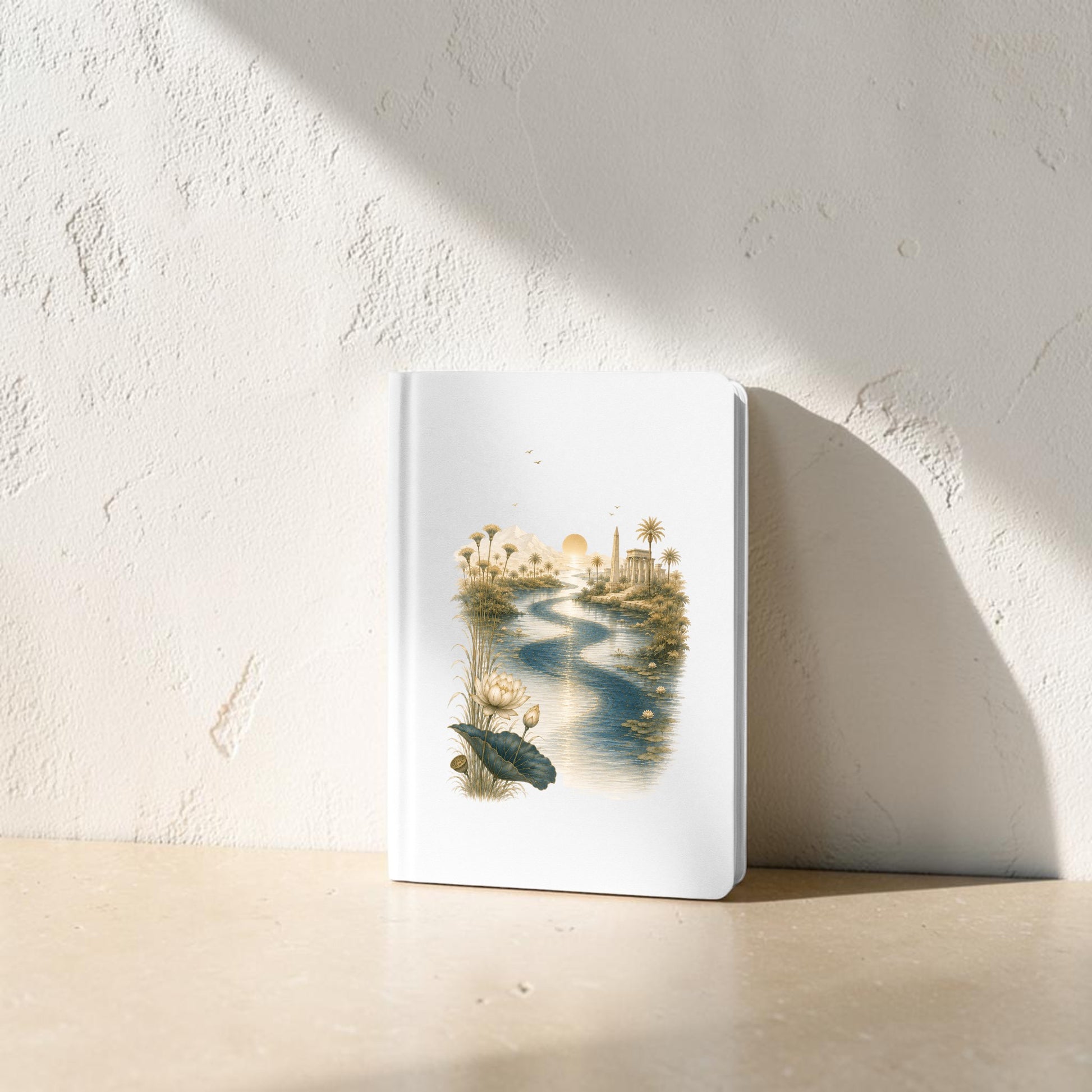 The Nile Path Journal | Ancient Egyptian Inspired Notebook by Mirabilia Boutique features a white cover adorned with a tranquil river and nature motif reminiscent of the serene Nile, accented with lotus and papyrus details. It rests on a beige surface against a textured wall, where sunlight bathes it in gentle shadows.