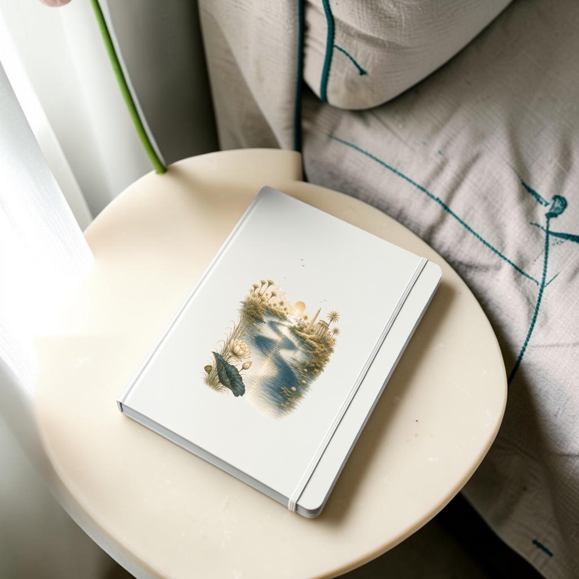 A Nile Path Journal | Ancient Egyptian Inspired Notebook by Mirabilia Boutique, featuring a white cover adorned with a scenic illustration of a river and trees that captures the tranquil spirit of the Nile Path, sits on a round beige table next to a bed draped with a patterned blanket.