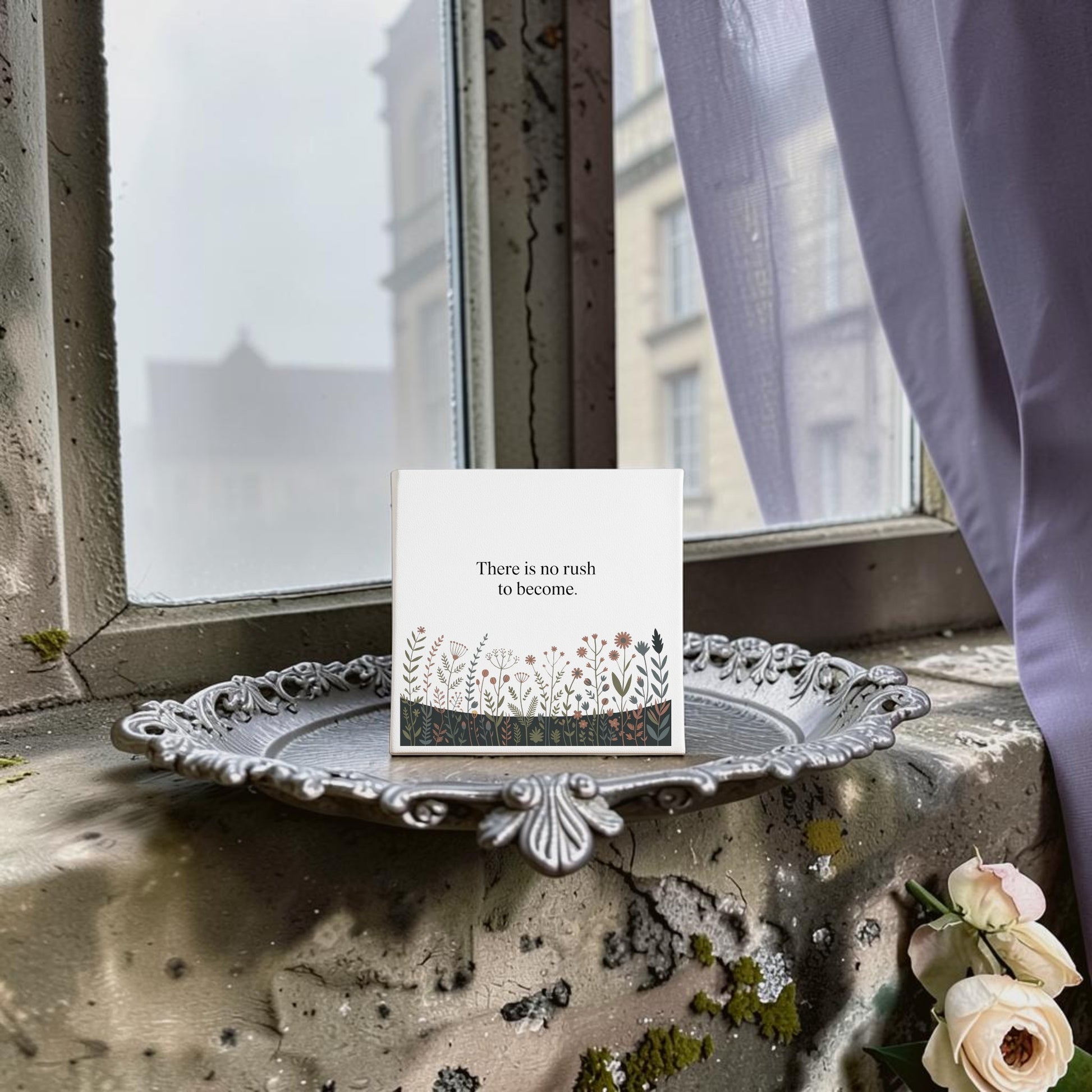 The Mirabilia Boutique "There Is No Rush to Become" Canvas Art features floral designs and motivational text, displayed on a mossy windowsill with pale roses—an inspiring decor piece or thoughtful gift, with city buildings visible beyond a sheer curtain.