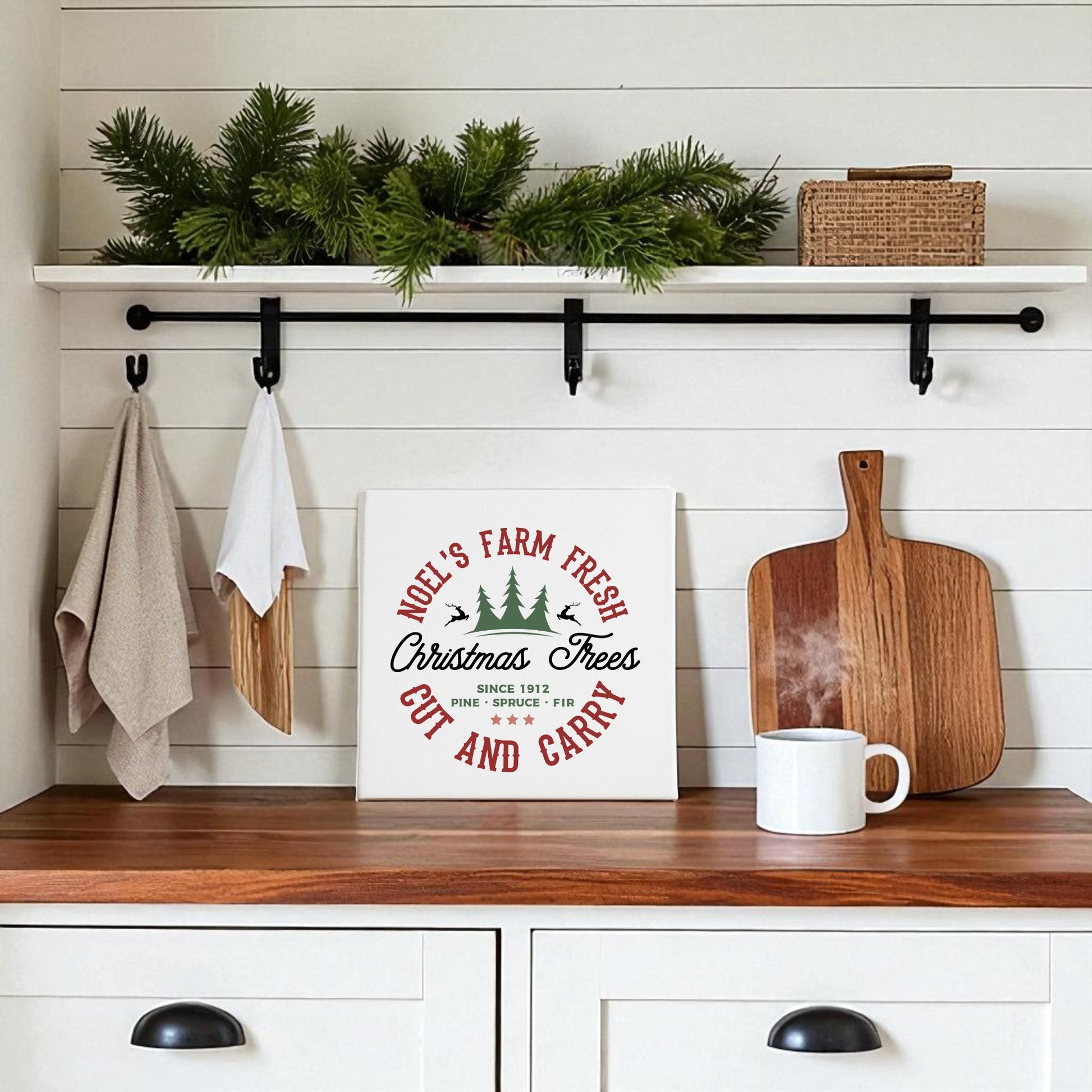 A wood kitchen counter holds a mug and cutting board. Above, festive decor includes greenery, a basket, towels, and the Mirabilia Boutique "Farm Fresh Christmas Tree" 8x8 canvas sign for a rustic holiday touch.