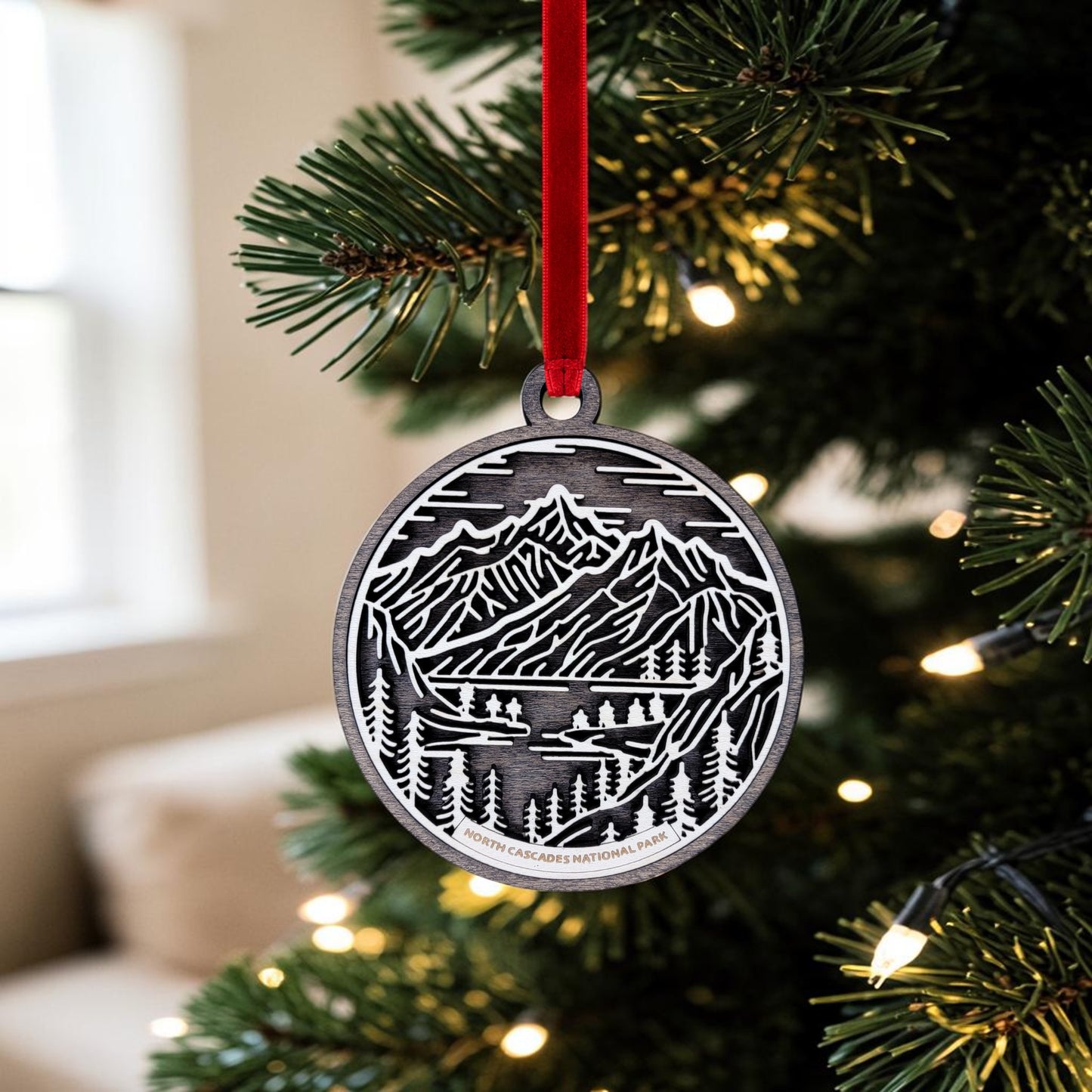 An oval, handcrafted North Cascades National Park Line Art Christmas Ornament by Mirabilia Boutique hangs from a Christmas tree branch on a red ribbon. Surrounded by twinkling white lights, this ornament brings the natural beauty of a mountain landscape to your holiday décor.