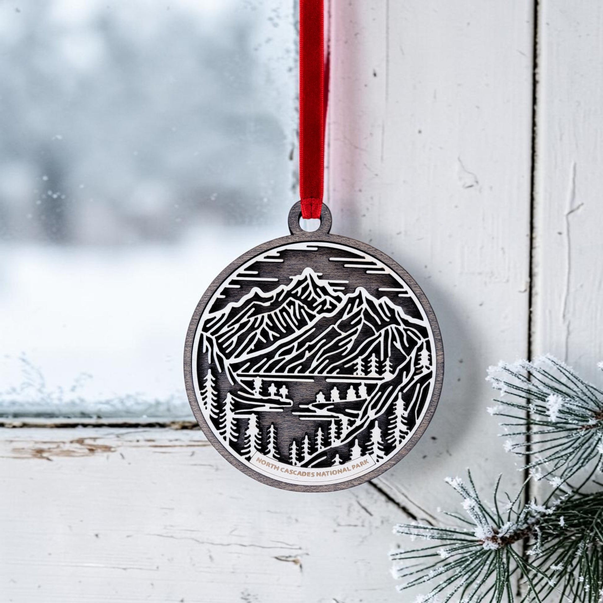 The North Cascades National Park Line Art Christmas Ornament by Mirabilia Boutique—a round, handcrafted keepsake featuring a mountain and forest scene—hangs from a red ribbon on a frosty window, with snowy pine needles in the corner and a wintry landscape visible outside.