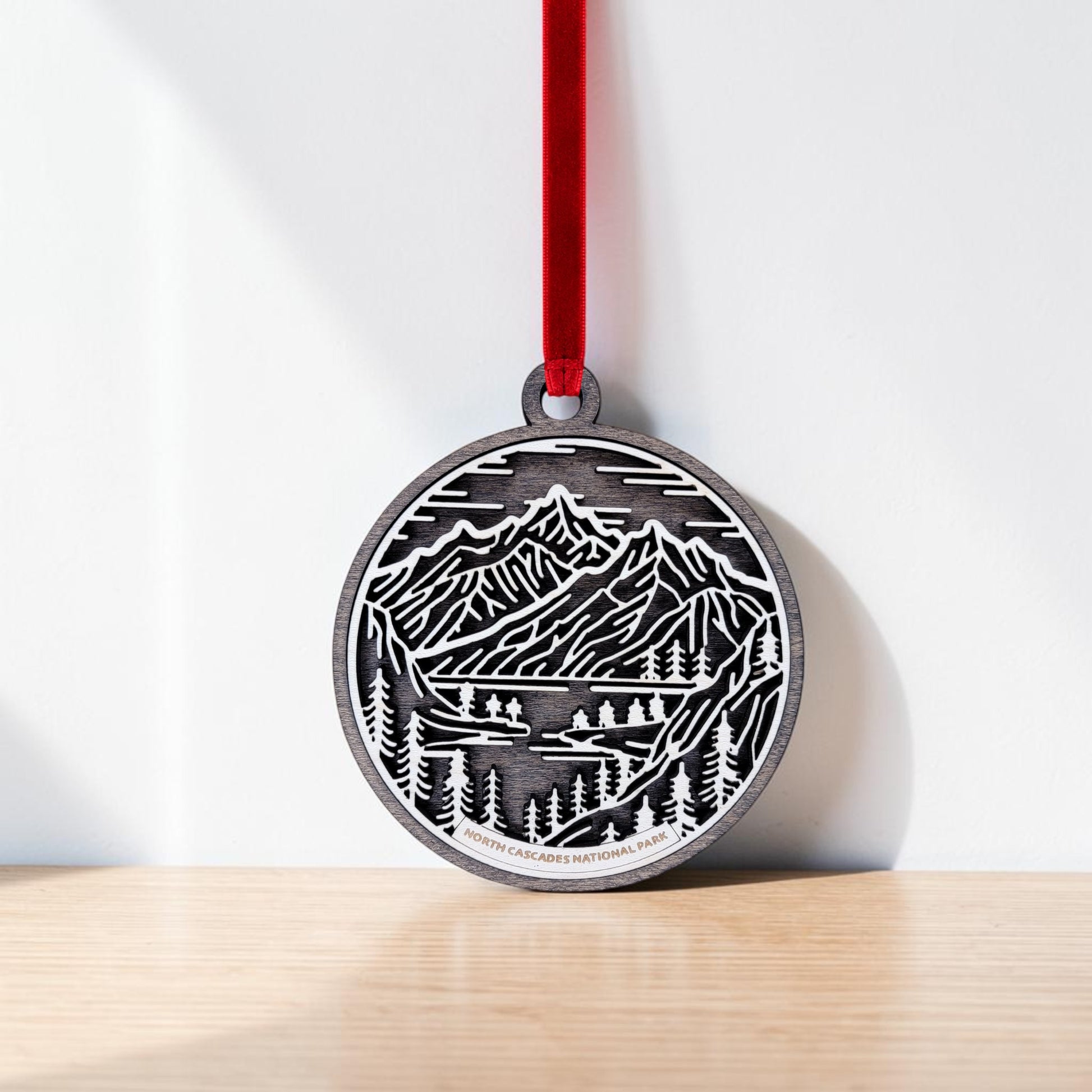 The North Cascades National Park Line Art Christmas Ornament by Mirabilia Boutique features a round wooden design engraved with a mountain and forest scene. Hanging from a red ribbon against a white wall and resting on a light wood surface, this handcrafted keepsake beautifully captures the essence of nature in elegant line art.