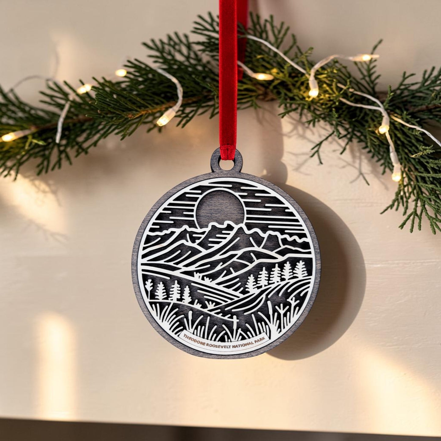 Adorn your holiday décor with the Theodore Roosevelt National Park Line Art Christmas Ornament by Mirabilia Boutique, featuring a mountain and landscape design. This unique badlands keepsake hangs from a red ribbon in front of green pine branches and string lights.