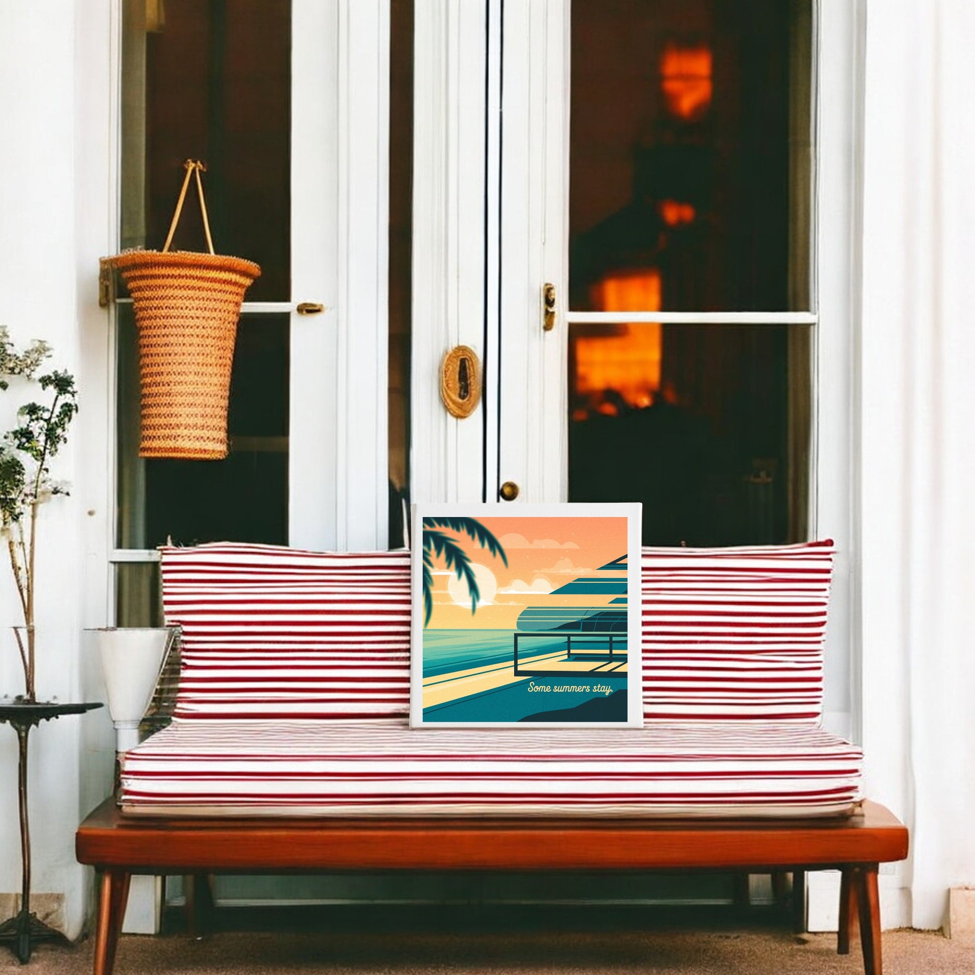 A striped red and white cushioned bench sits before white French doors. The "Some Summers Stay Canvas – Retro Coastal Sunset Art" by Mirabilia Boutique rests on the bench, while a woven basket hangs near a potted plant.