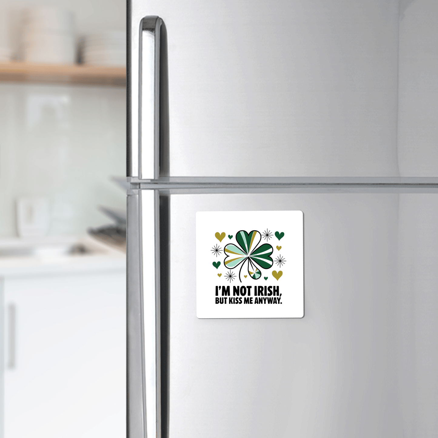 A Mirabilia Boutique "Kiss Me Anyway Funny St. Patrick’s Day Magnet" with a shamrock and green hearts decorates a refrigerator; kitchen shelves and dishes are visible in the background.