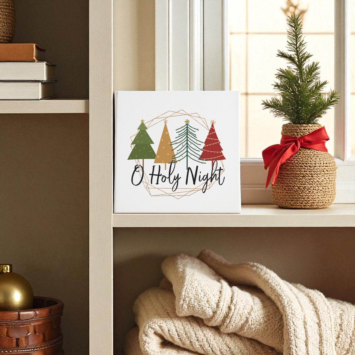 A shelf styled with the Mirabilia Boutique O Holy Night Canvas Sign (8x8), festive potted pine, books, gold ornament, and folded blankets creates easy minimalist Christmas decor.
