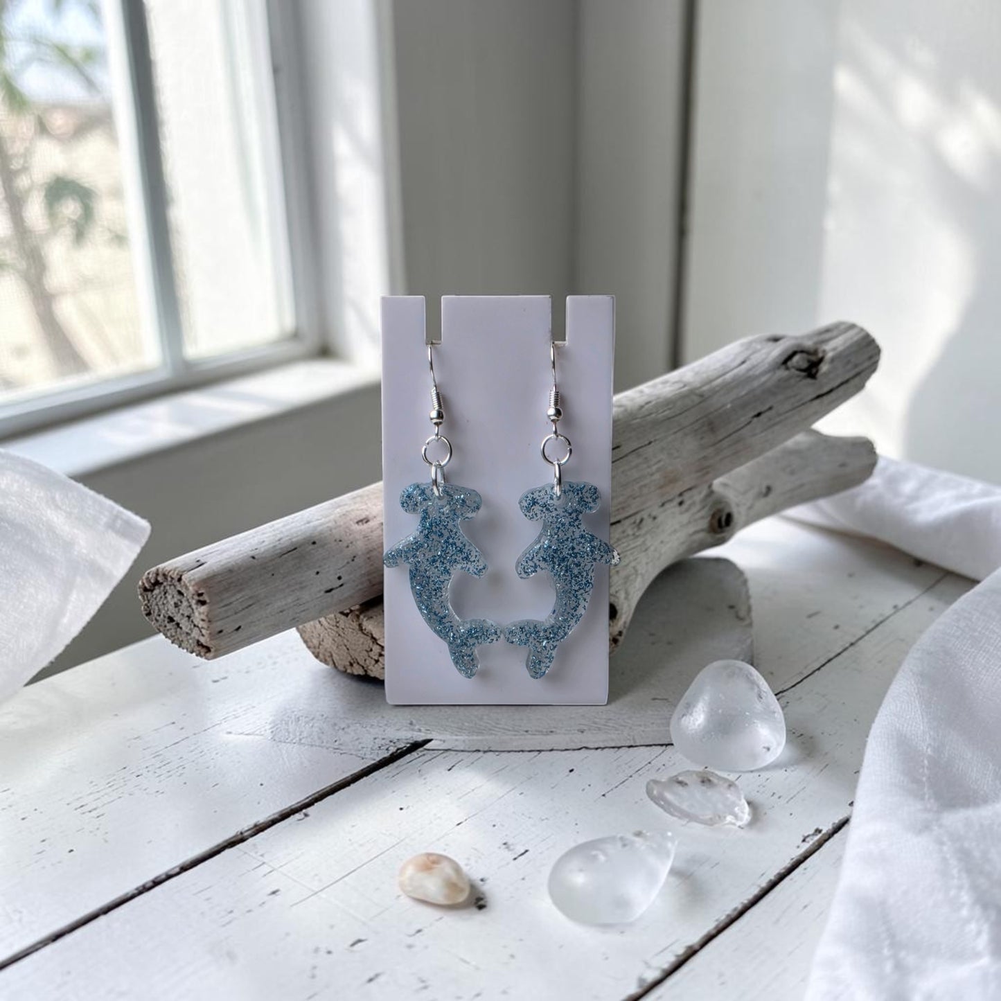 Blue Glitter Hammerhead Shark Earrings by Mirabilia Boutique hang from a white card on driftwood, surrounded by sea glass and shells on a sunlit white wooden surface, creating an ocean-inspired jewelry display.