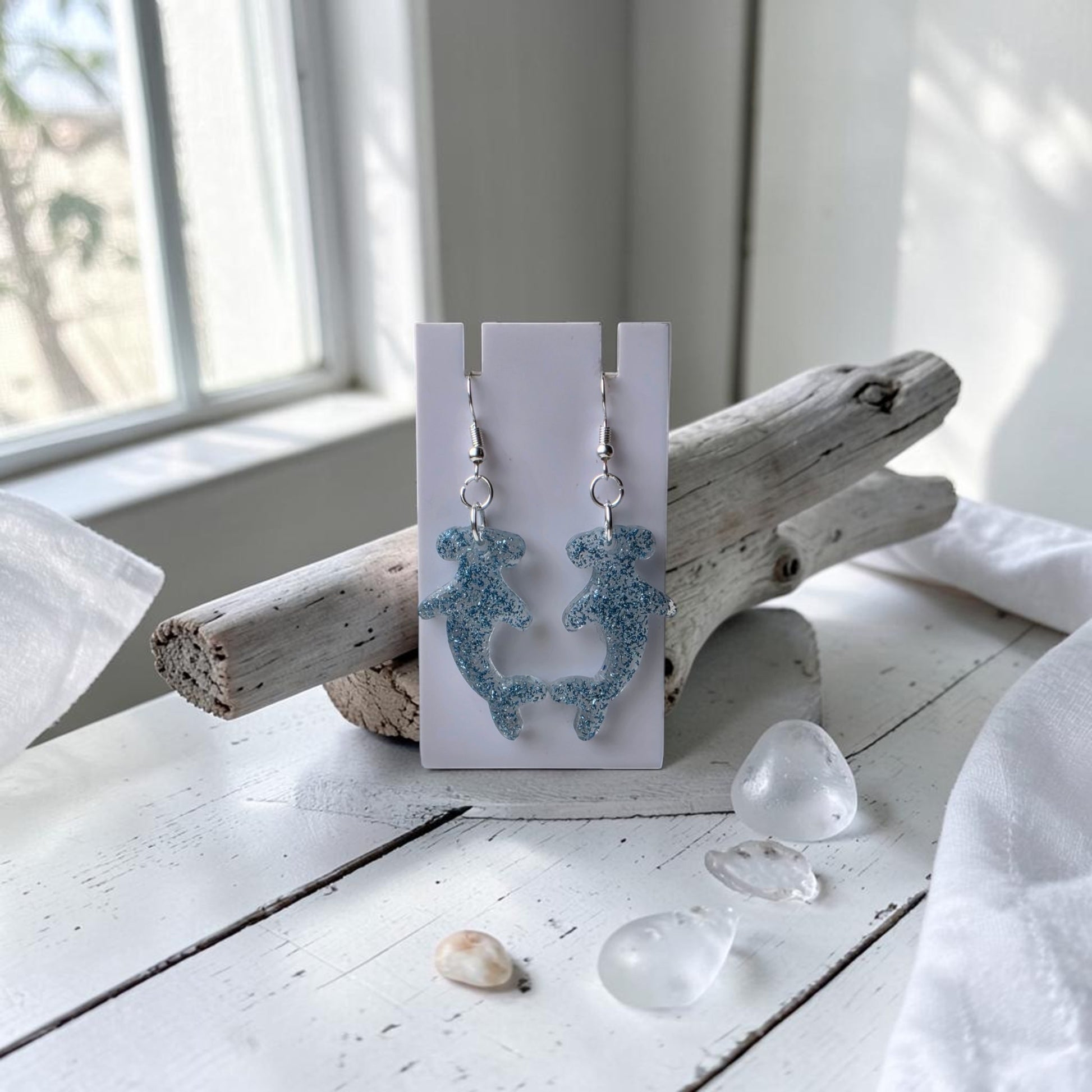 Blue Glitter Hammerhead Shark Earrings by Mirabilia Boutique hang from a white card on driftwood, surrounded by sea glass and shells on a sunlit white wooden surface, creating an ocean-inspired jewelry display.