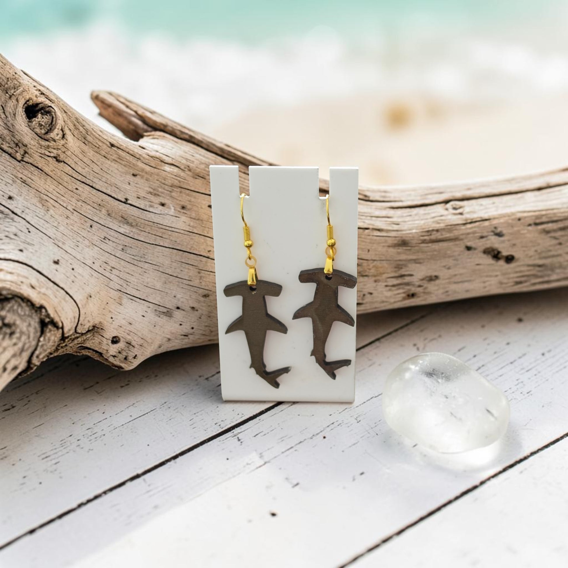 Hammerhead Shark Earrings | Lasercut Walnut Wood by Mirabilia Boutique hang on a white card, set against driftwood with a frosted glass pebble and soft beach background—an eye-catching marine life accessory with gold hooks.