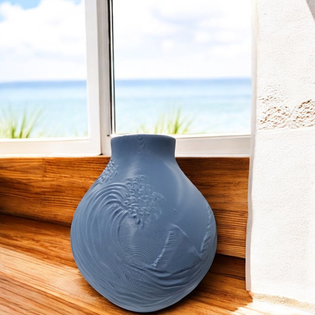 The Stone Blue Coastal Wave Vase by Mirabilia Boutique, featuring embossed patterns, rests on a wooden windowsill with sheer curtains, while an ocean and sky view outside enhances the serene coastal decor.
