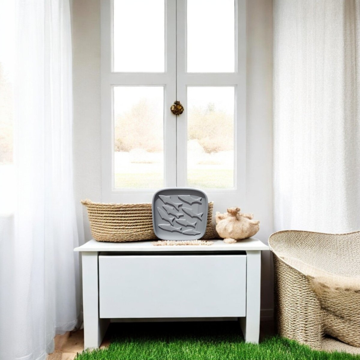 A white table holds the Mirabilia Boutique Coastal Fish Decorative Tray and woven baskets by bright windows with sheer curtains; nearby is a wicker chair, all set atop green grass flooring.