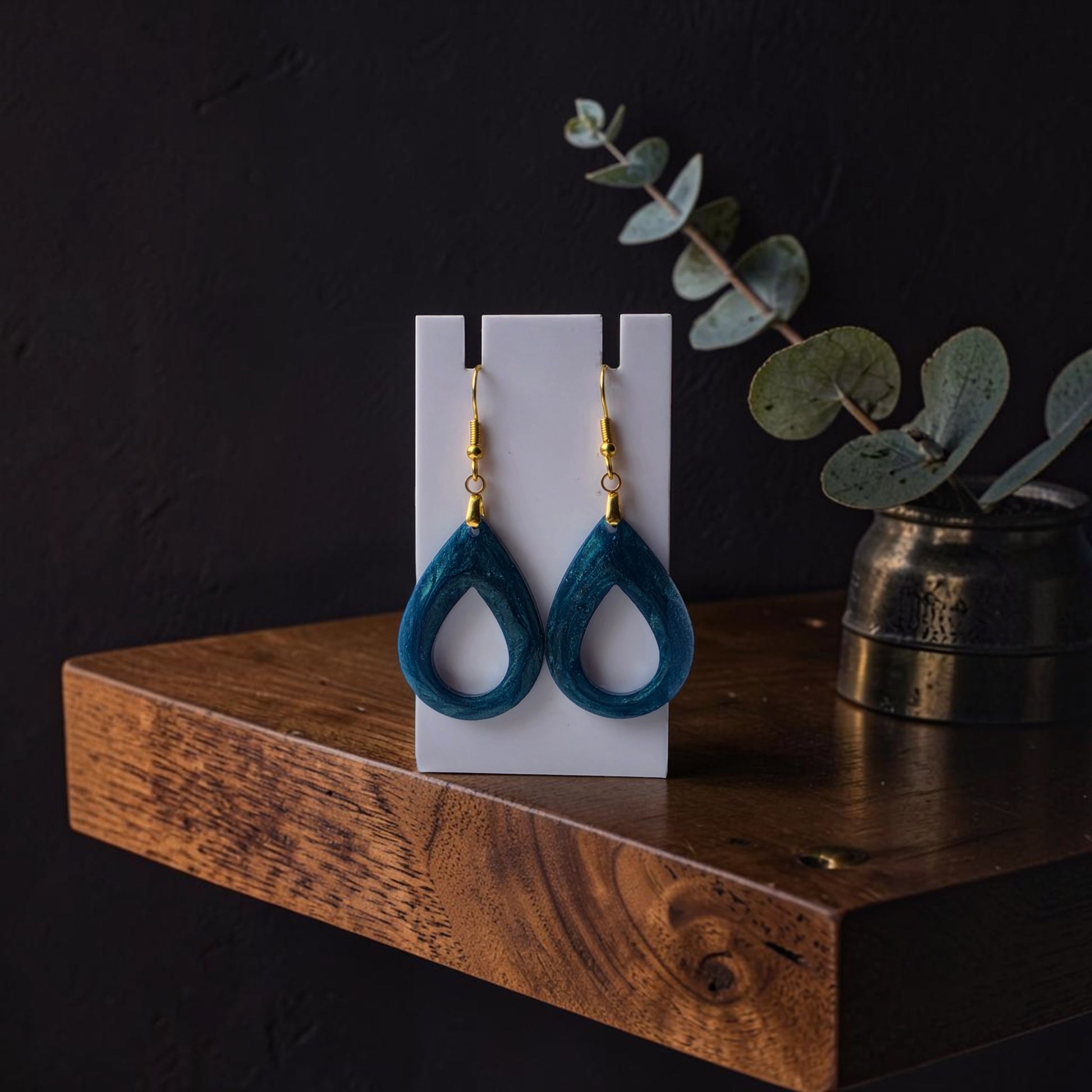 The Mermaid Dust Teardrop Earrings by Mirabilia Boutique feature handcrafted blue teardrop stones with gold hooks, displayed on a white stand atop a wooden shelf with eucalyptus branches in the background.