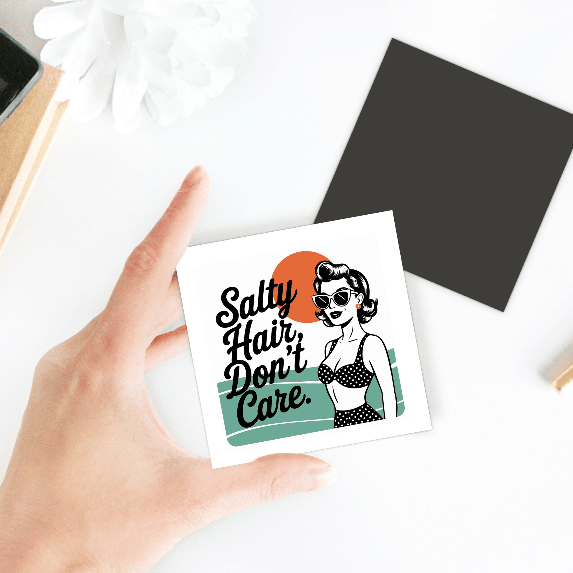 A hand holds the Mirabilia Boutique "Salty Hair Don’t Care Magnet – Retro Beach Quote," featuring a woman in sunglasses and polka-dot swimsuit, ideal for beach house decor. A white flower and black square sit on a white desk behind.