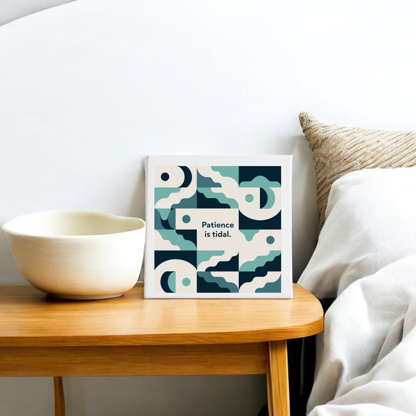 The Patience Is Tidal Canvas by Mirabilia Boutique, a calm coastal wall art print, rests on a wooden side table beside a white bowl, with a beige textured pillow on the sofa or bed in the background.