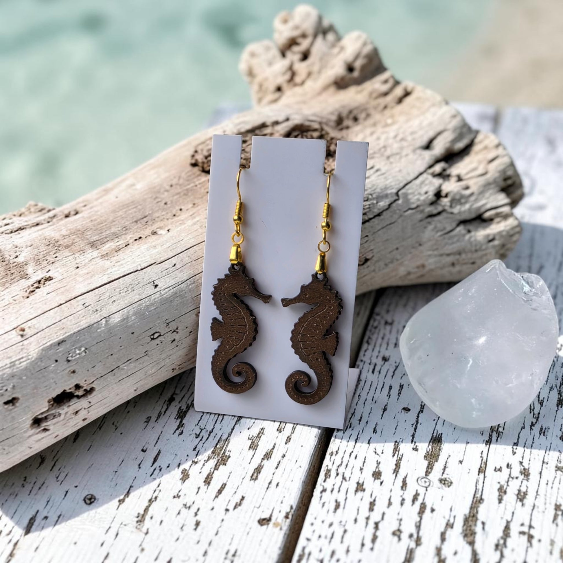 A pair of Seahorse Earrings | Lasercut Walnut Wood by Mirabilia Boutique hang on a display card atop weathered white wood with driftwood and a clear crystal; blurred water shimmers gently in the background.