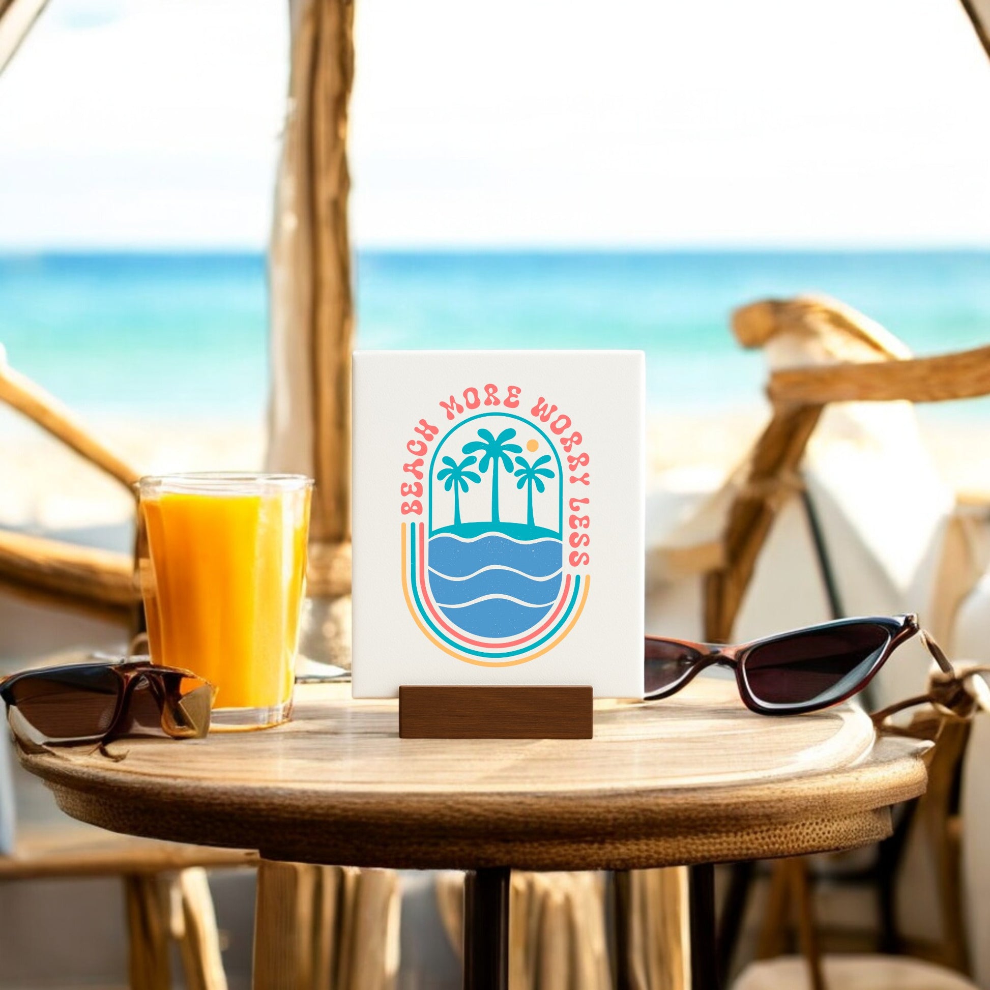 A table by the beach displays Mirabilia Boutique's Beach More Worry Less Retro Summer Sign with palm tree and wave graphics, plus sunglasses and orange juice—perfect summer decor for an ocean view or as a seasonal gift.