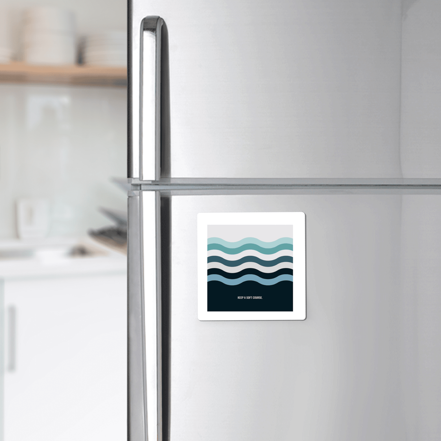 The Keep a Soft Course Magnet by Mirabilia Boutique, featuring a minimalist ocean-inspired wavy blue design, is displayed on a stainless steel refrigerator, bringing subtle coastal decor to this modern kitchen.