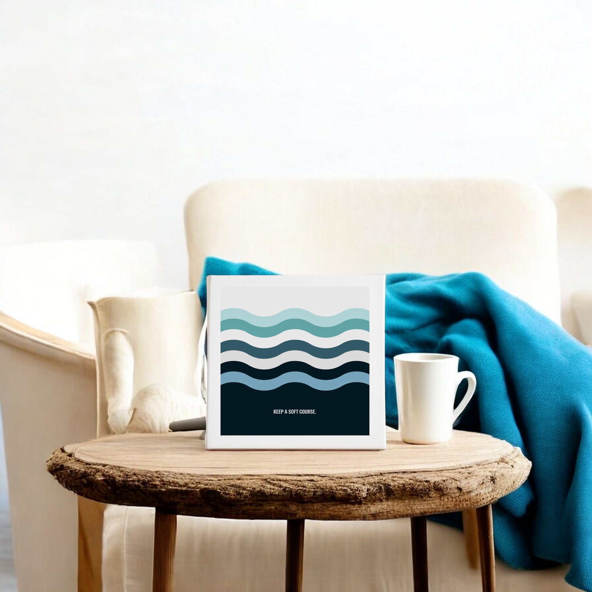 The Mirabilia Boutique "Keep a Soft Course" 8x8 canvas sign featuring minimalist blue waves rests on a rustic wooden table beside a white mug, with a cream armchair and blue blanket adding calm to the decor.