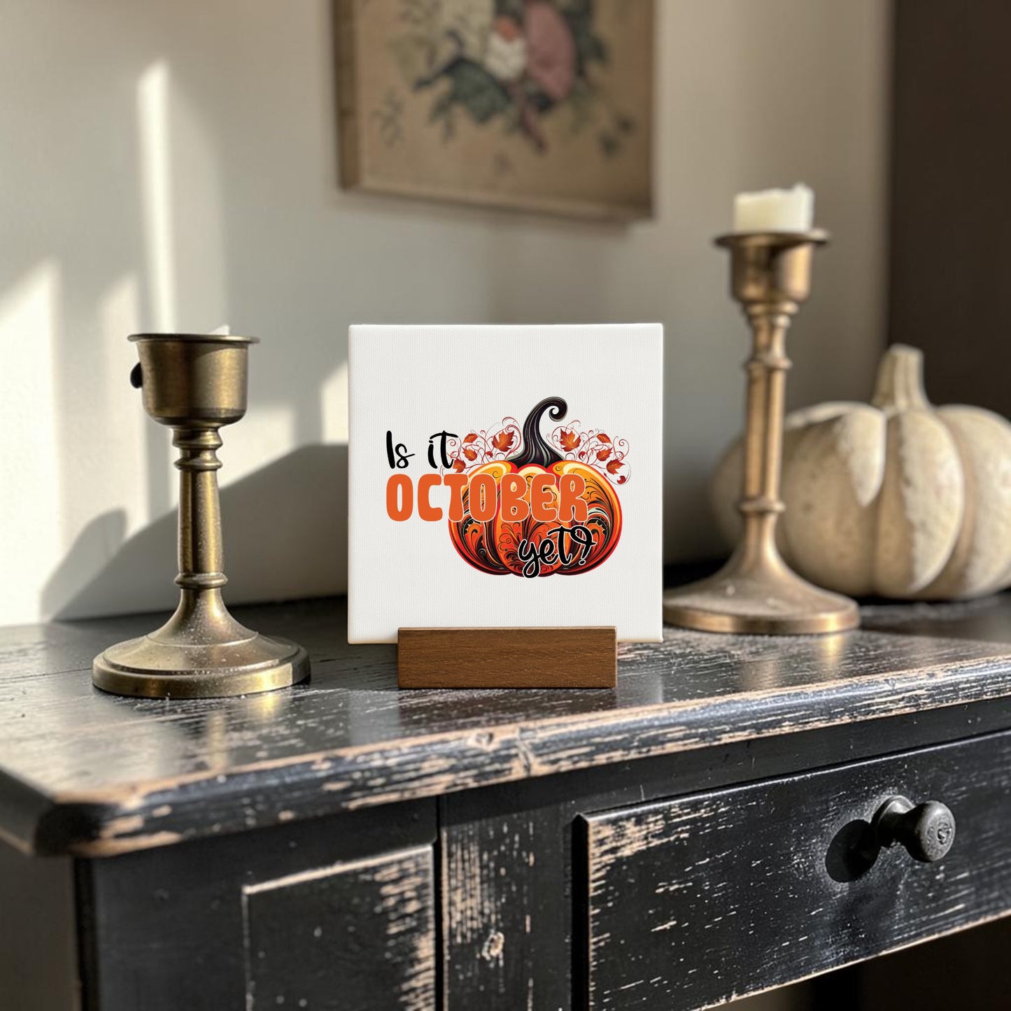 The "Is It October Yet? Mini Sign" by Mirabilia Boutique sits atop a wooden stand, surrounded by candle holders and a white pumpkin, creating cozy fall decor perfect for October.