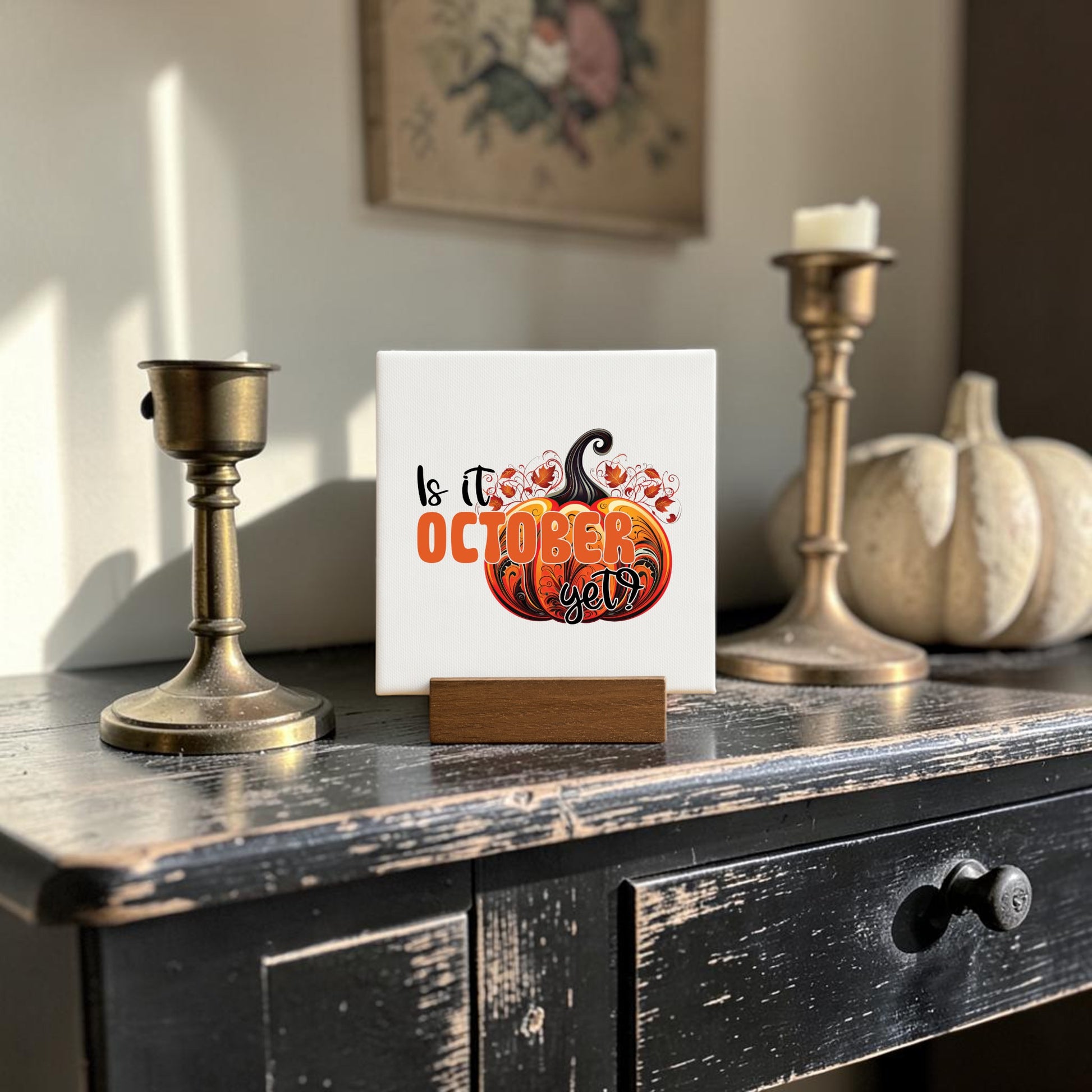 The "Is It October Yet? Mini Sign" by Mirabilia Boutique sits atop a wooden stand, surrounded by candle holders and a white pumpkin, creating cozy fall decor perfect for October.