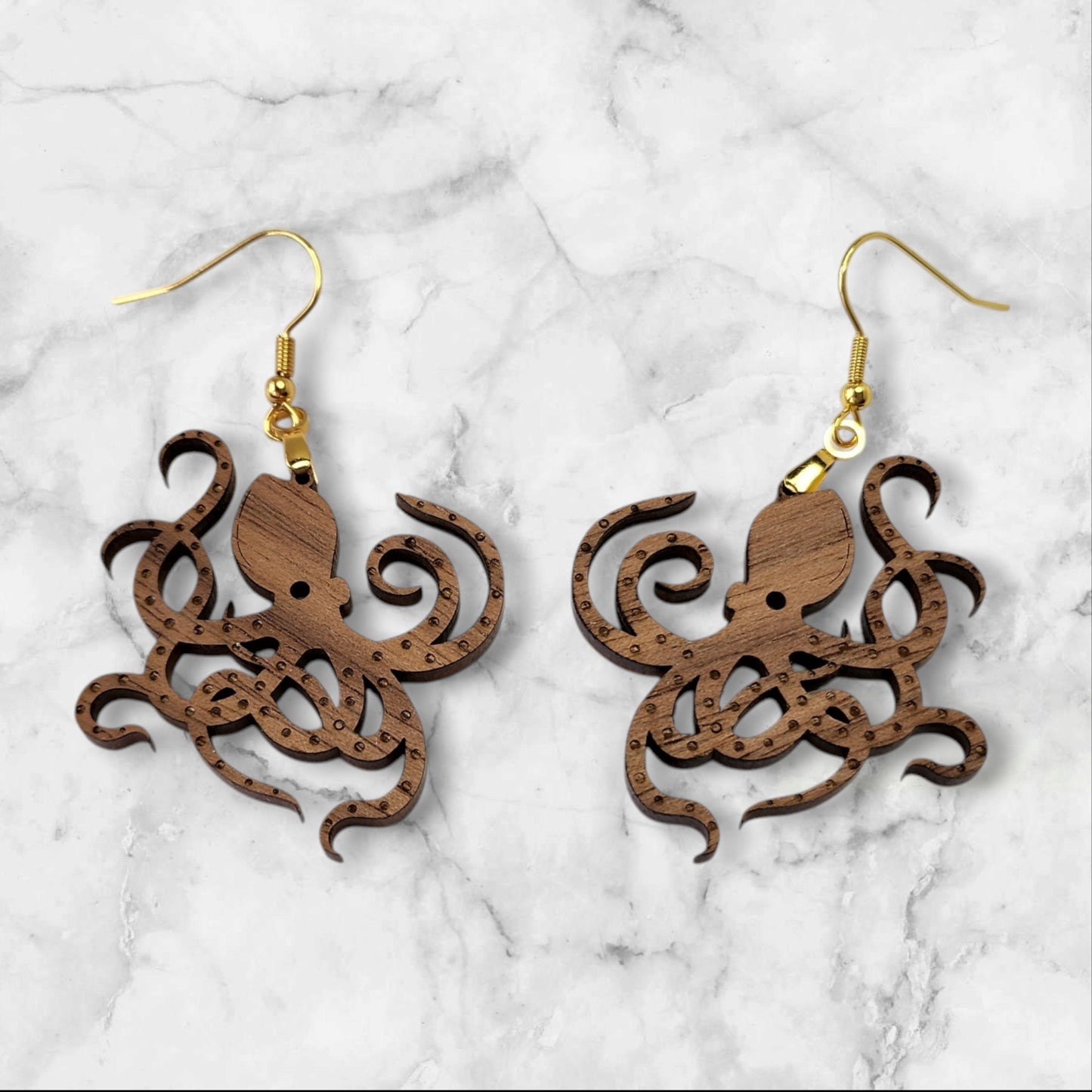 The Octopus Earrings from Mirabilia Boutique feature intricate laser-cut designs in walnut wood, hanging elegantly on gold hooks against a marble background.