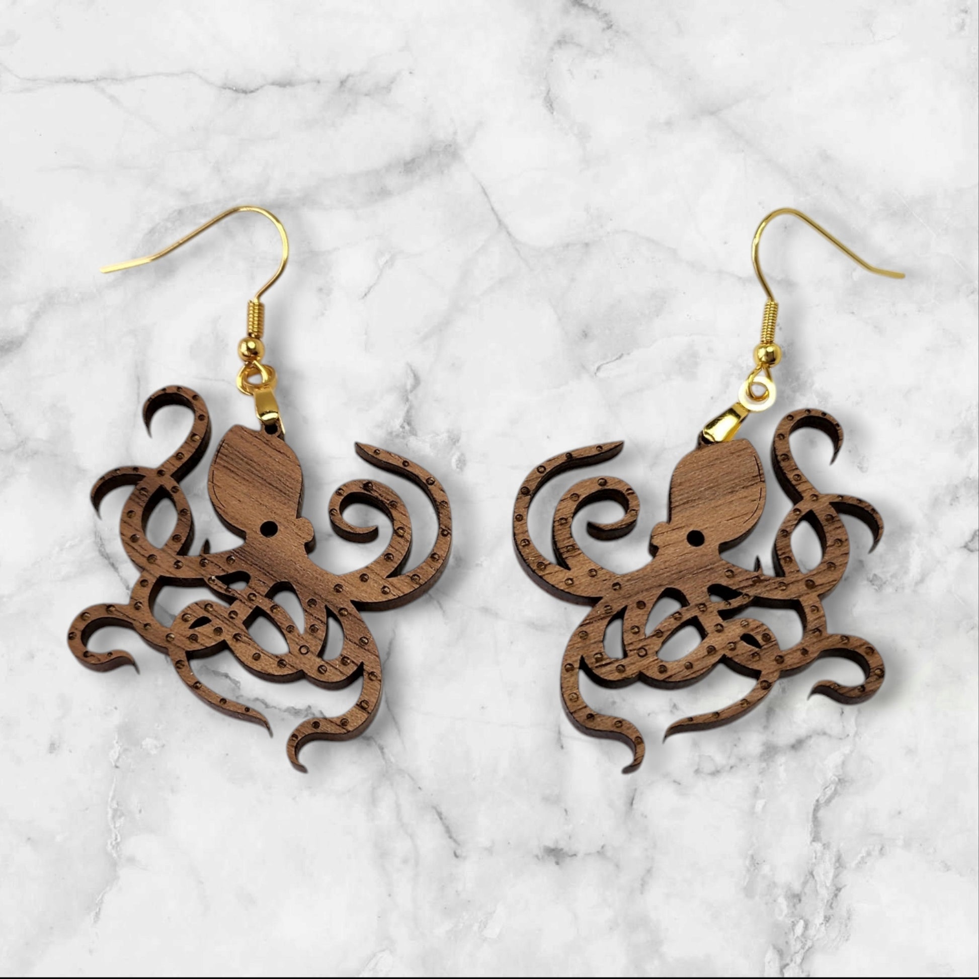 The Octopus Earrings from Mirabilia Boutique feature intricate laser-cut designs in walnut wood, hanging elegantly on gold hooks against a marble background.