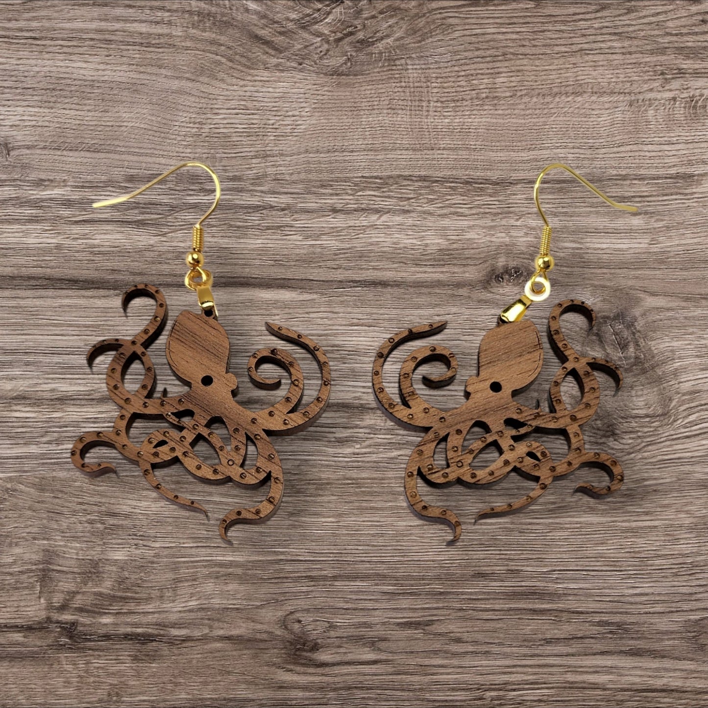 Mirabilia Boutique presents the Octopus Earrings: intricately laser-cut, walnut wood earrings featuring detailed tentacles and gold hooks, elegantly set on a textured wooden surface.