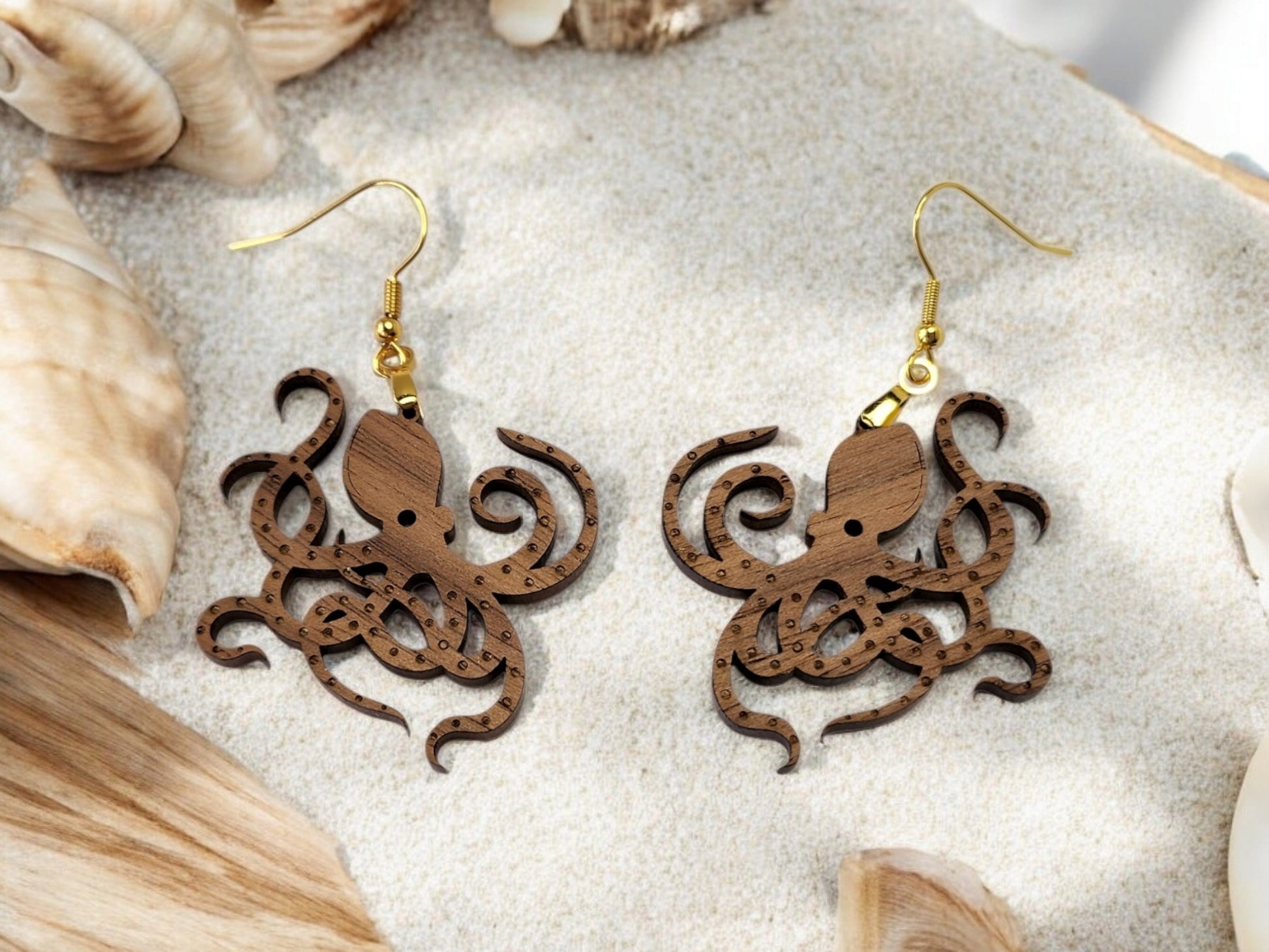 Mirabilia Boutique's Octopus Earrings, crafted from walnut wood with golden hooks, are beautifully displayed on sand with seashells and driftwood, giving a beach aesthetic. Their detailed laser cut design enhances the elegant seaside vibe.