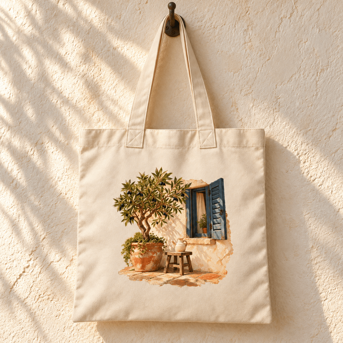 The Olive and Light Tote Bag by Mirabilia Boutique hangs on a hook, featuring an illustration of an olive tree in a pot, a small wooden stool, and an open window with blue shutters.