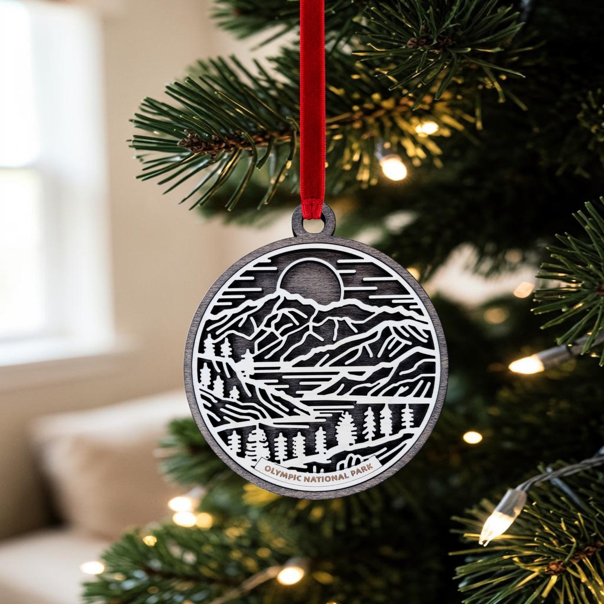 A metal line art keepsake from Mirabilia Boutique, the Olympic National Park Line Art Christmas Ornament, features an engraved mountain and forest scene and hangs from a red ribbon on a Christmas tree decorated with white lights. The ornament reads "Olympic National Park" at the bottom.