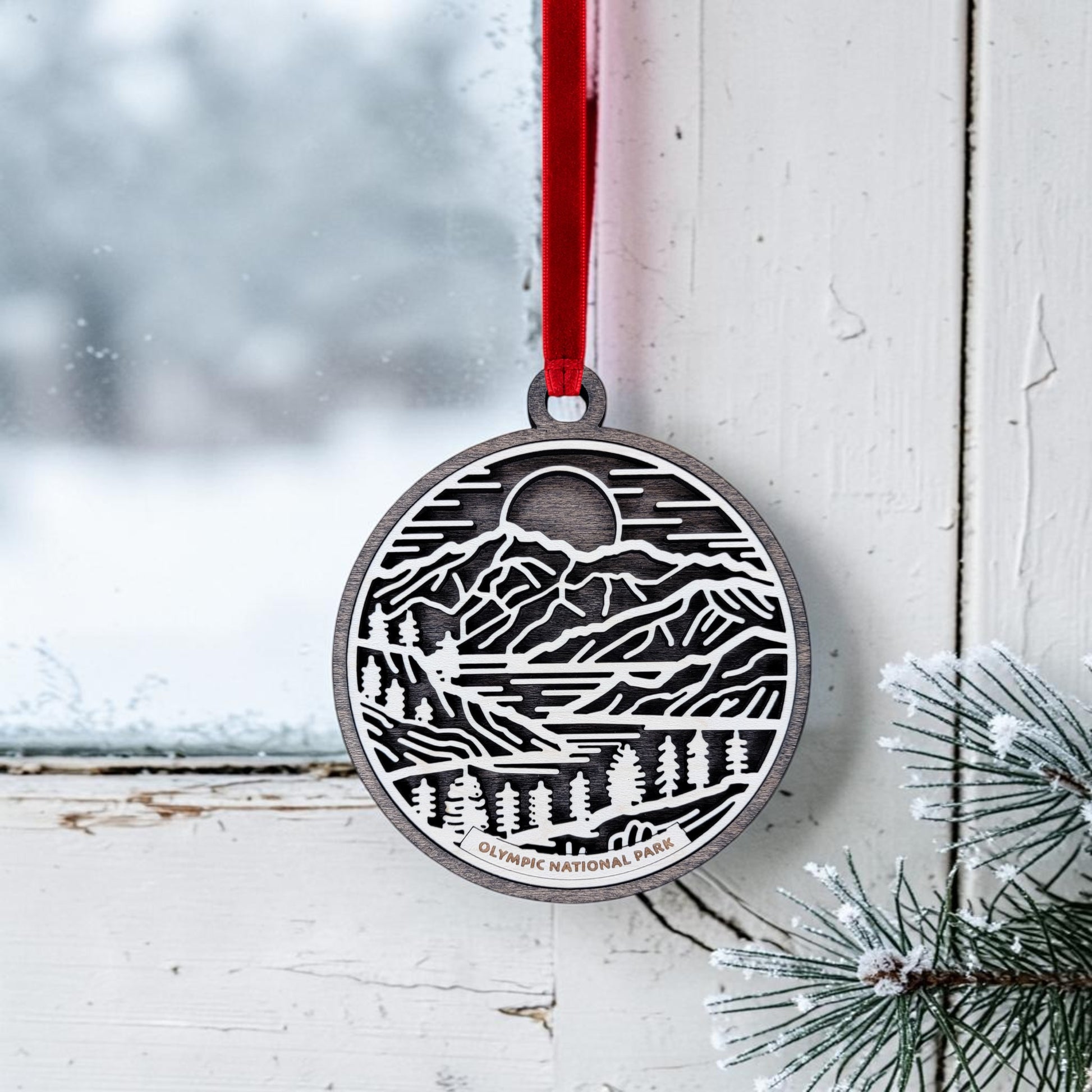A handcrafted Olympic National Park Line Art Christmas Ornament by Mirabilia Boutique hangs by a red ribbon on a frosty window, featuring mountains, trees, and a moon. Pine branches peek into the bottom right corner. The ornament is inscribed with “Olympic National Park,” making it an ideal keepsake for nature lovers.