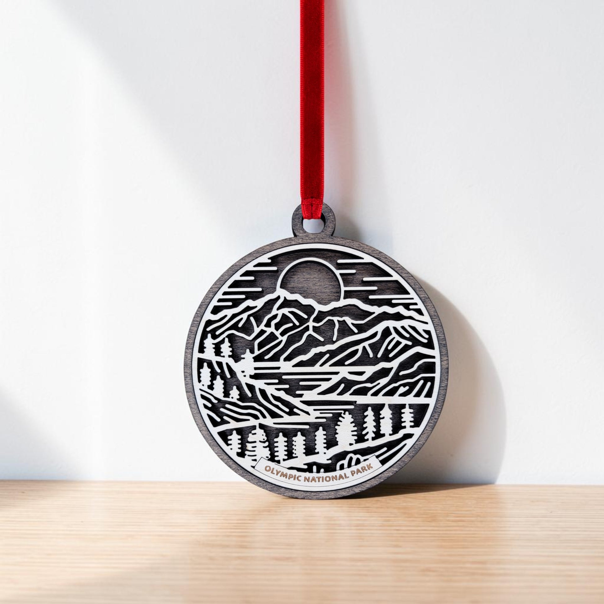 A round, handcrafted wooden ornament from Mirabilia Boutique, the Olympic National Park Line Art Christmas Ornament, hangs from a red ribbon against a white wall. Featuring delicate line art of mountains, trees, and the sun, this keepsake is inscribed with "Olympic National Park" at the base.
