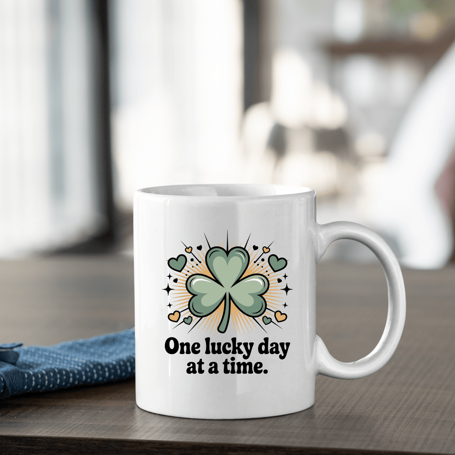The Mirabilia Boutique "One Lucky Day at a Time" Shamrock Ceramic Mug features a green four-leaf clover with yellow accents, displayed on a wooden table next to a folded blue cloth.