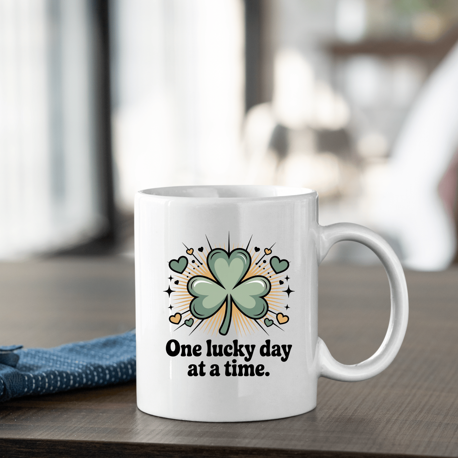 The Mirabilia Boutique "One Lucky Day at a Time" Shamrock Ceramic Mug features a green four-leaf clover with yellow accents, displayed on a wooden table next to a folded blue cloth.