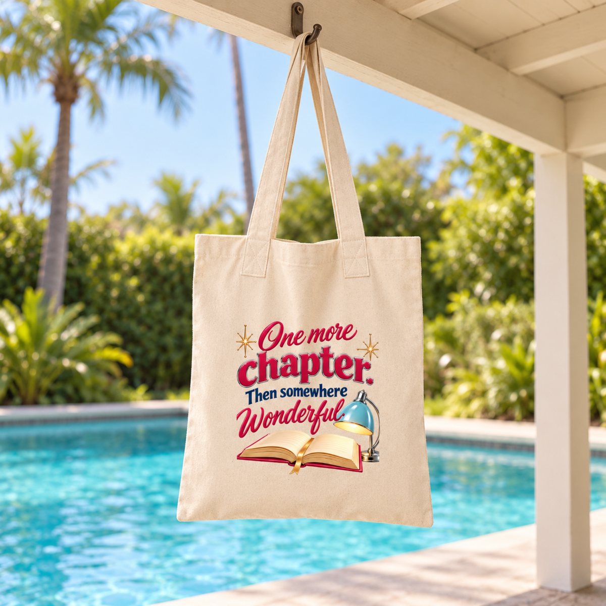 A cream-colored One More Chapter Book Lover Tote | Chrome & Starlight featuring an illustration of an open book and a desk lamp, with the text “One more chapter. Then somewhere wonderful” printed in red and blue.