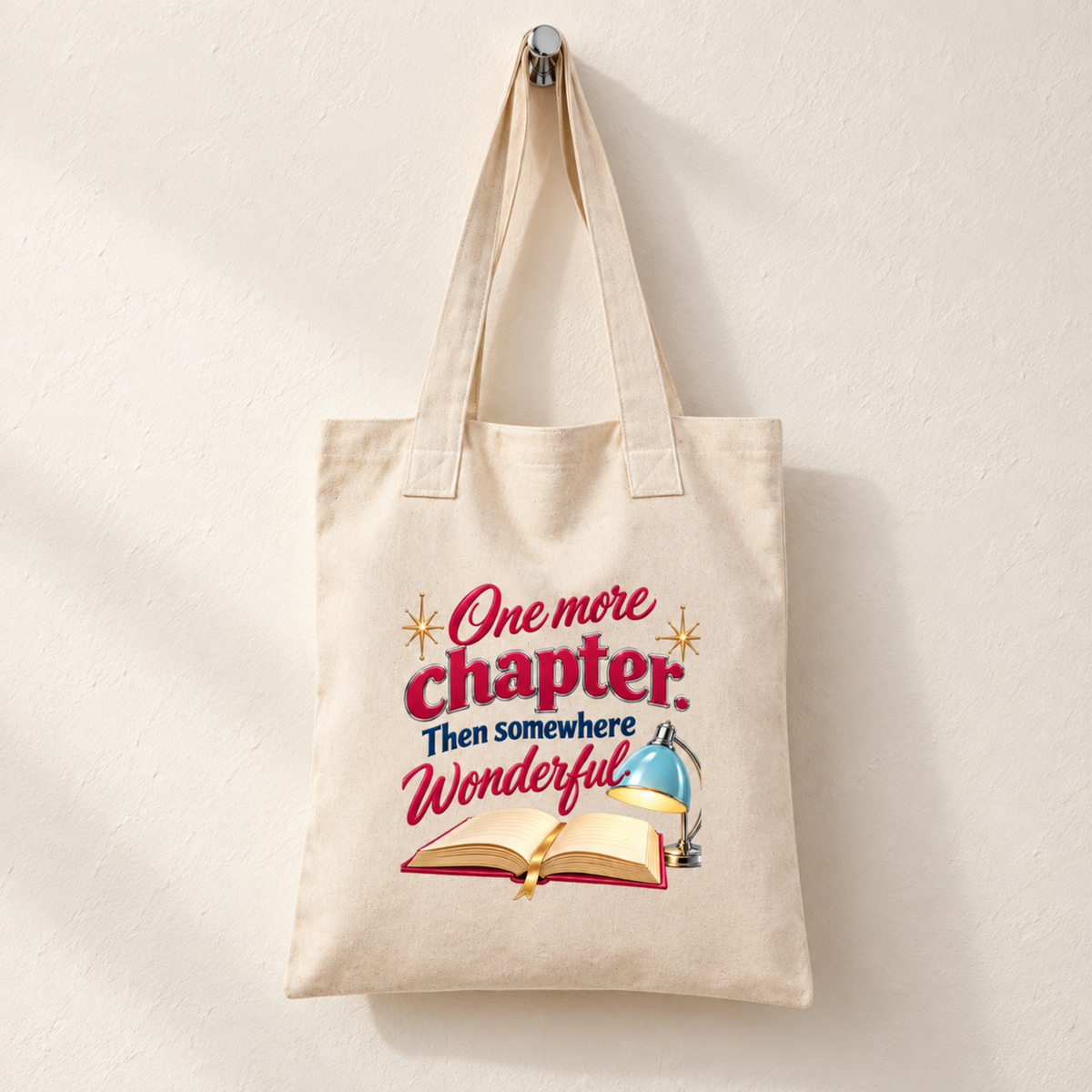 One More Chapter Book Lover Tote by Mirabilia Boutique hanging on a wall hook, featuring a print of an open book, a desk lamp, and the text “One more chapter. Then somewhere wonderful.”.