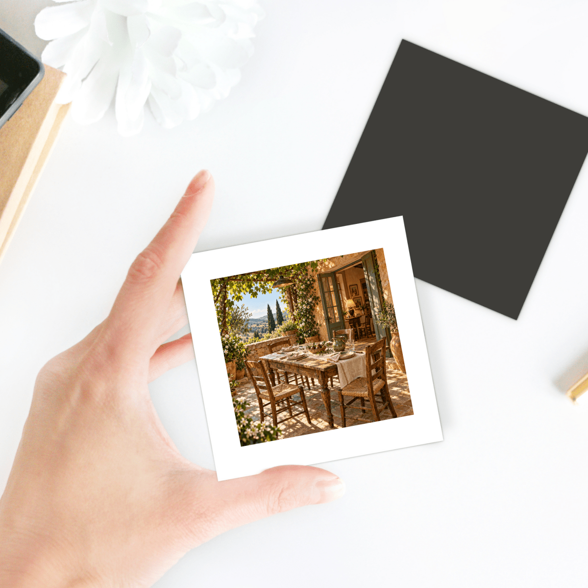A hand holds a square One More Place Tuscan Art Magnet | Ochre & Cypress, featuring a printed image of a dining table set on a sunlit stone patio.