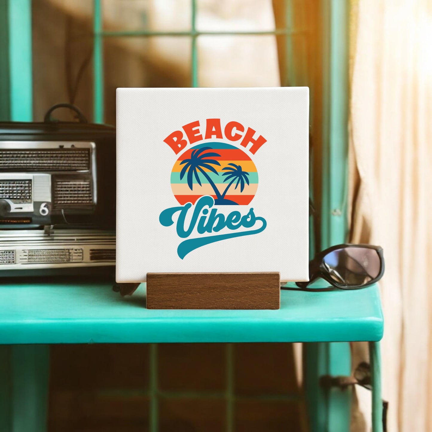 The Mirabilia Boutique Beach Vibes Sunset Canvas – Bold Tropical Mini Sign displays a sunset, palm trees, and "Beach Vibes," standing on a turquoise shelf by a vintage radio and sunglasses with sunlight through beige-curtained windows.